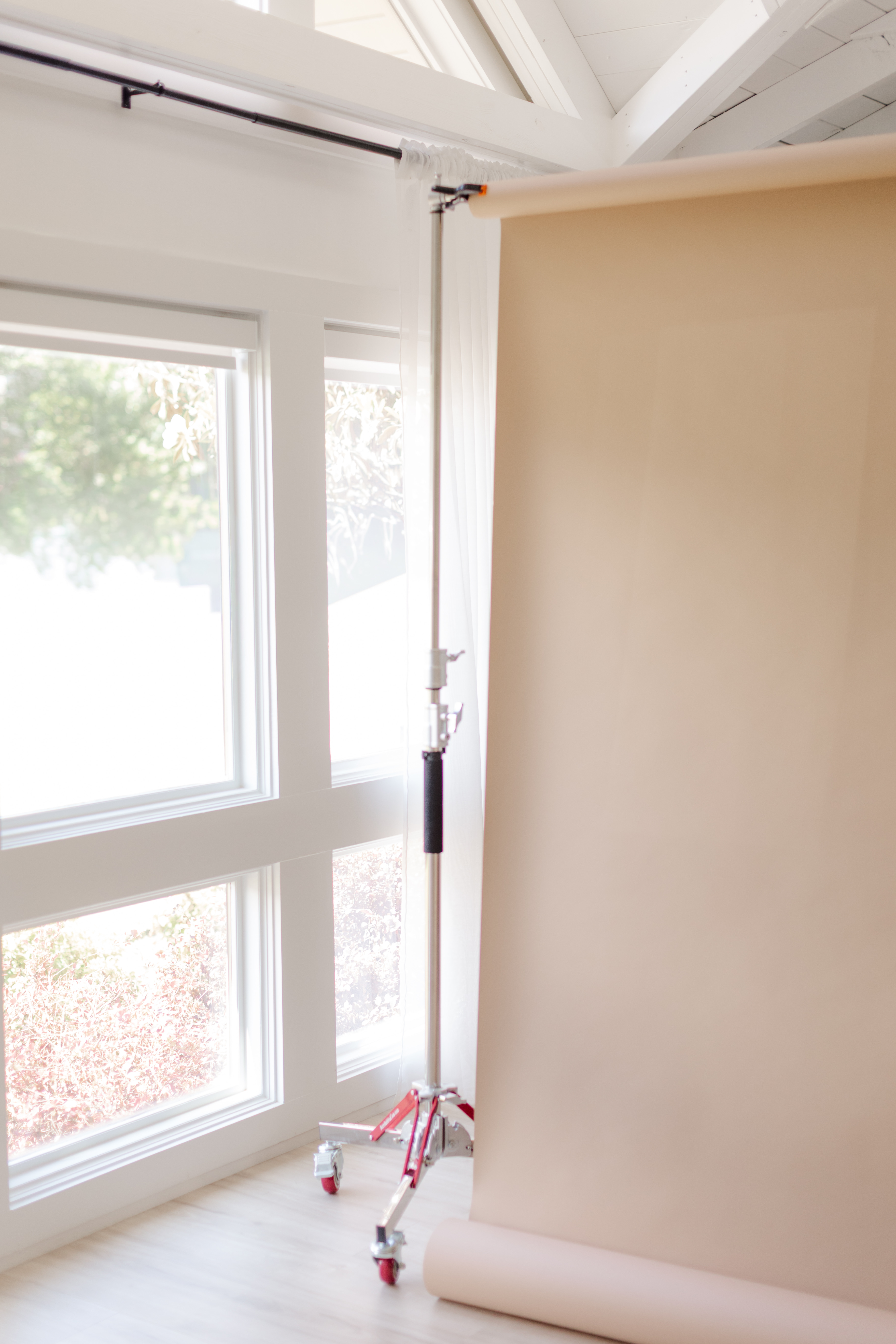 Photography backdrop roll with beige paper hanging on a stand near a window in a bright room.