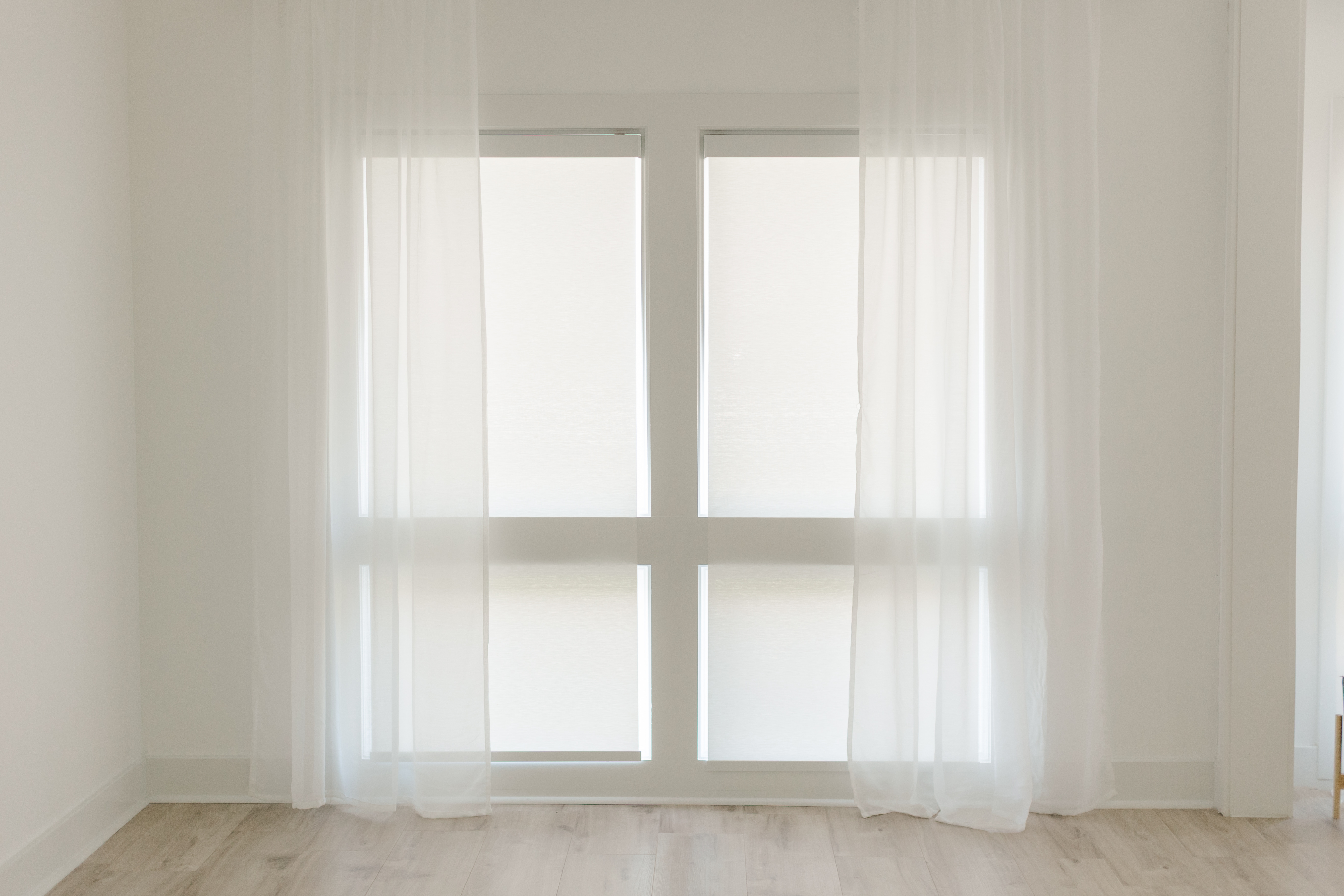 Large window with sheer white curtains and light wooden flooring in a bright empty room.