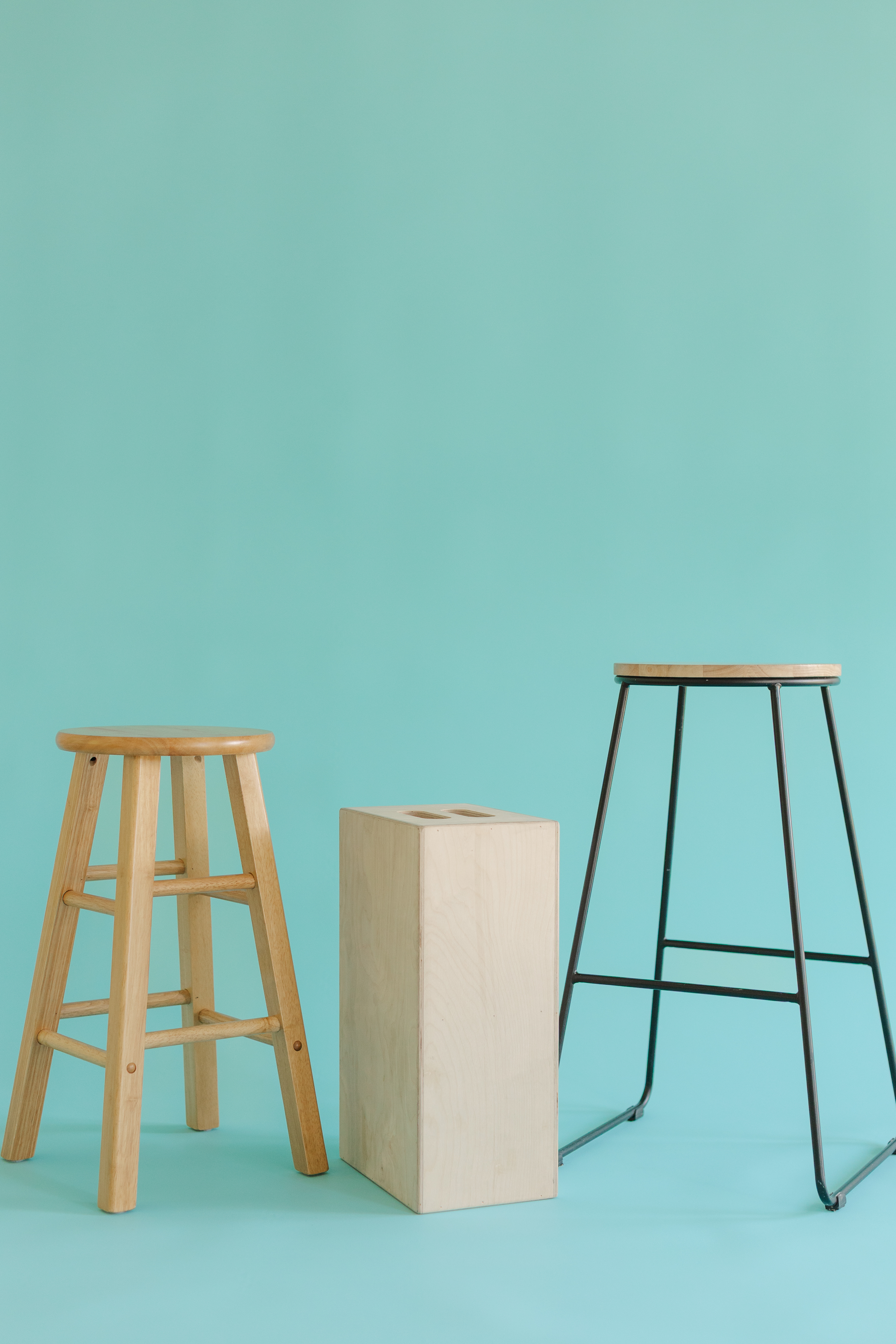 Three stools on a turquoise background: wooden round seat, rectangular wooden block, and metal frame stool.