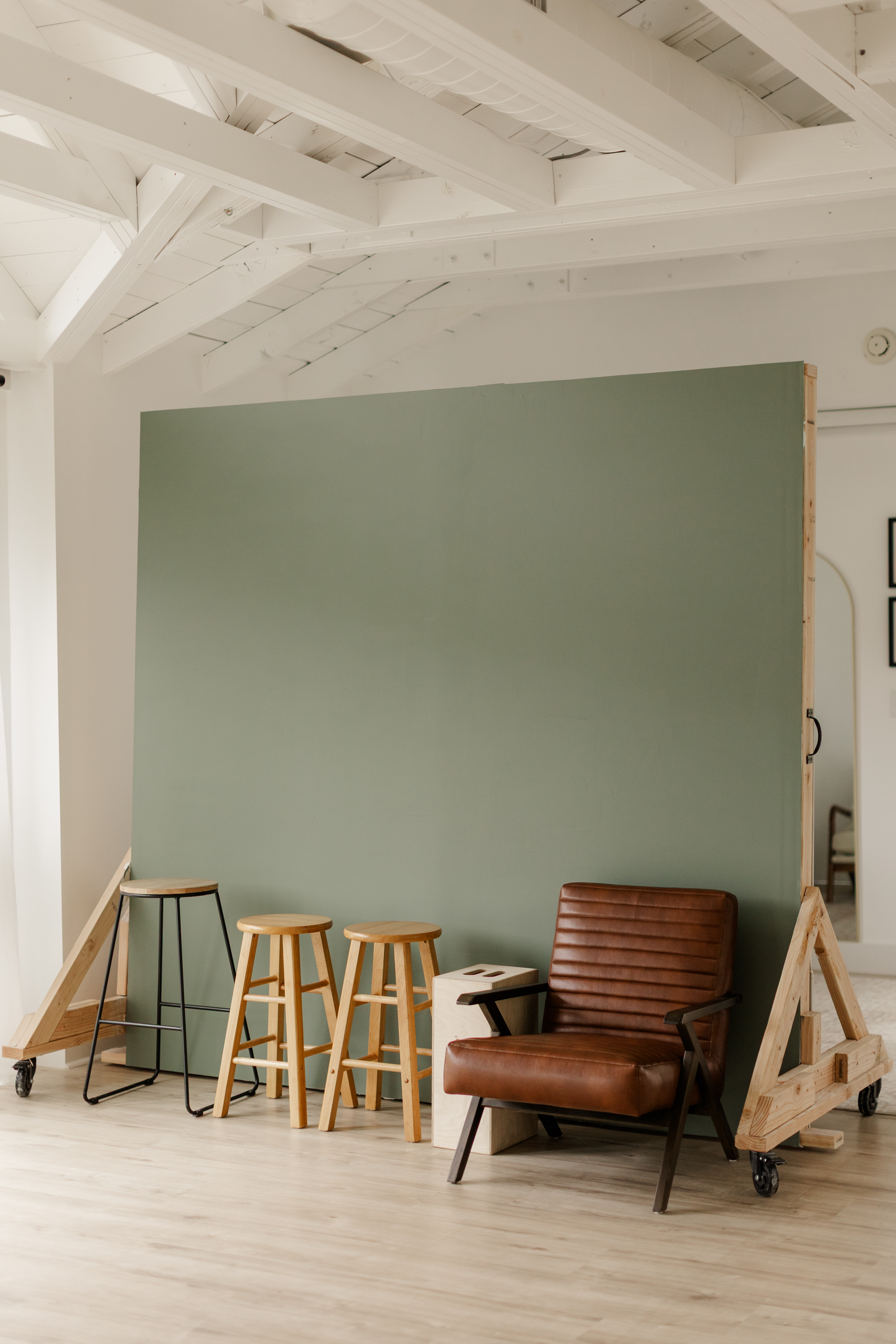 Modern indoor setup with a green divider, three wooden stools, and a brown leather armchair on wood floor.
