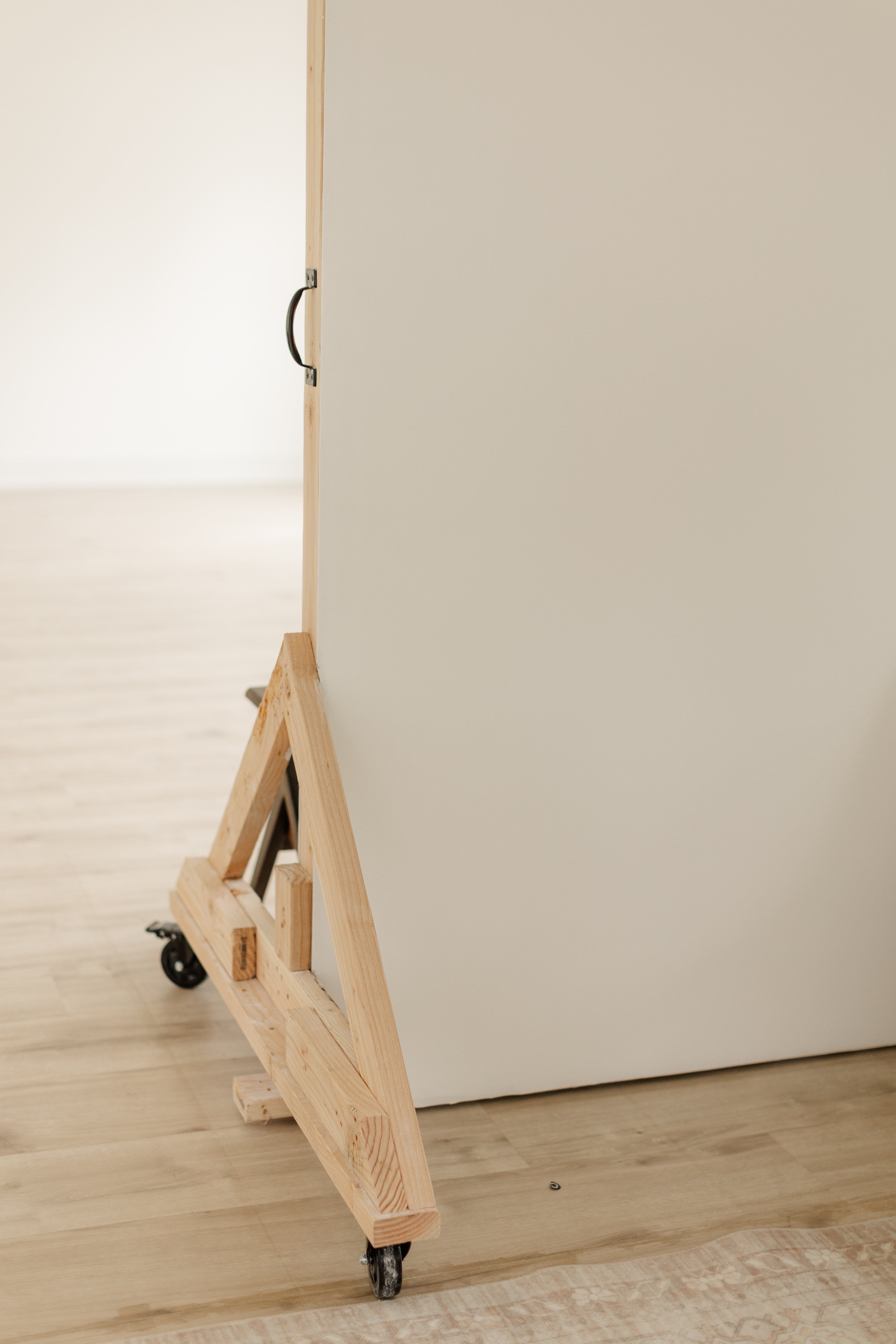 Close-up of a wooden easel with wheels holding a large blank white canvas on a wooden floor.