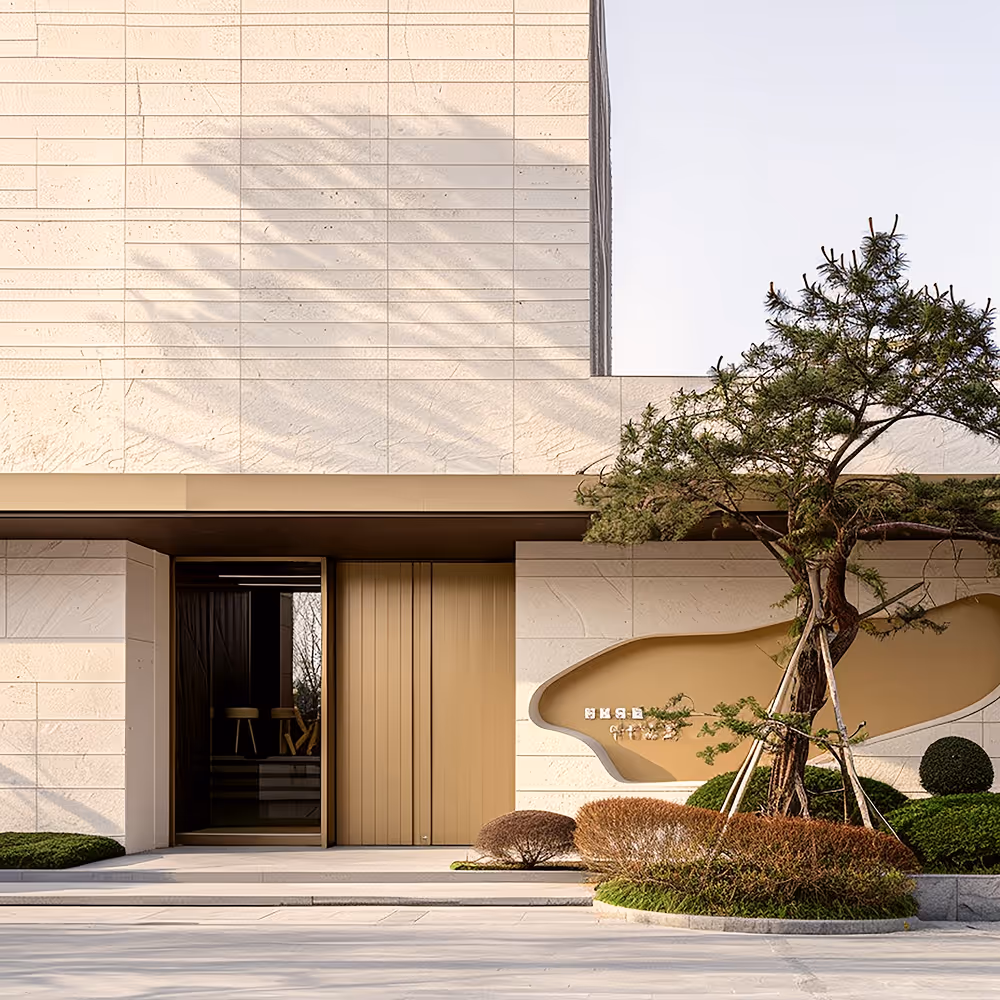 Modern building entrance with large beige panels, vertical golden door, and a landscaped garden featuring a curved tree supported by wooden stakes.