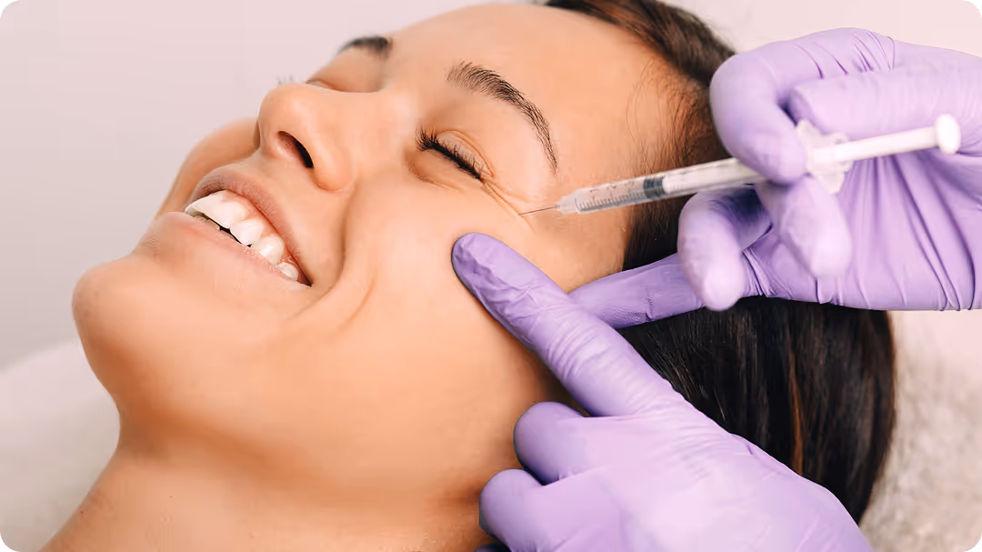 Close-up of a smiling woman receiving a cosmetic injection near her eye from a gloved professional.