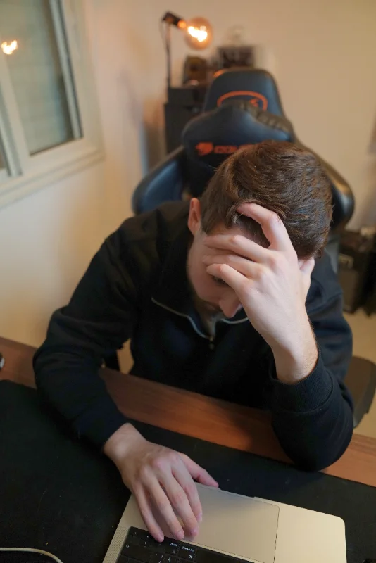 Man sitting at a desk with a laptop, holding his forehead with one hand appearing stressed or deep in thought.