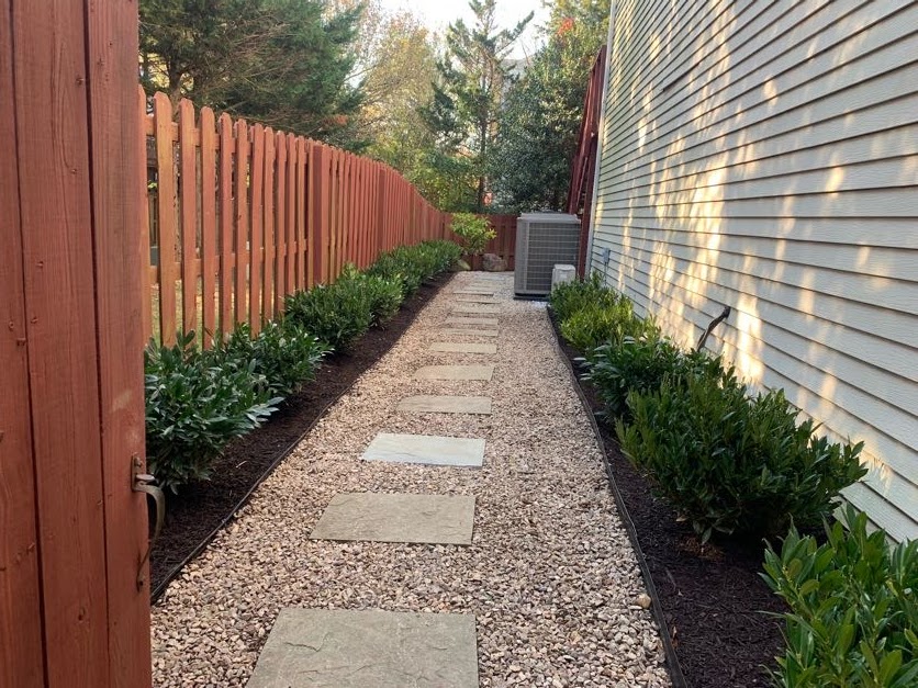 A landscaped side yard walkway featuring a path of light-colored gravel and rectangular stepping stones, bordered by garden beds of dark mulch and green shrubs, located between a wooden fence and a house with light-colored siding.