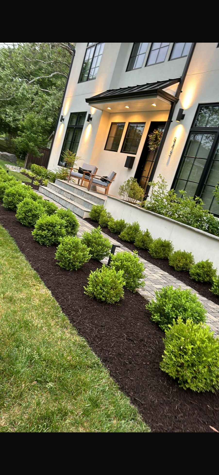 A landscaped side yard walkway featuring a path of light-colored gravel and rectangular stepping stones, bordered by garden beds of dark mulch and green shrubs, located between a wooden fence and a house with light-colored siding.