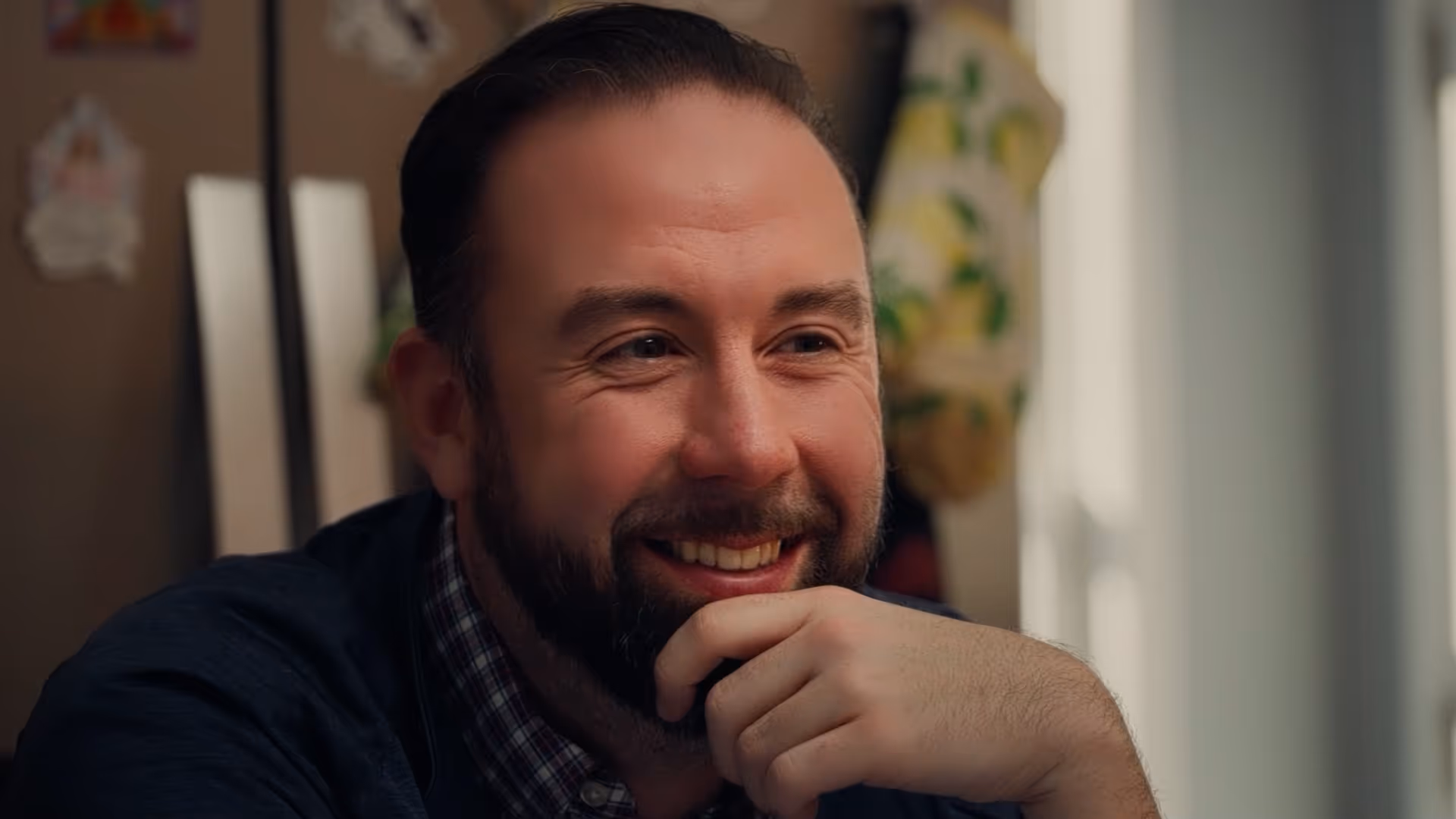 Smiling man with beard resting chin on hand, looking off-camera in warmly lit room.