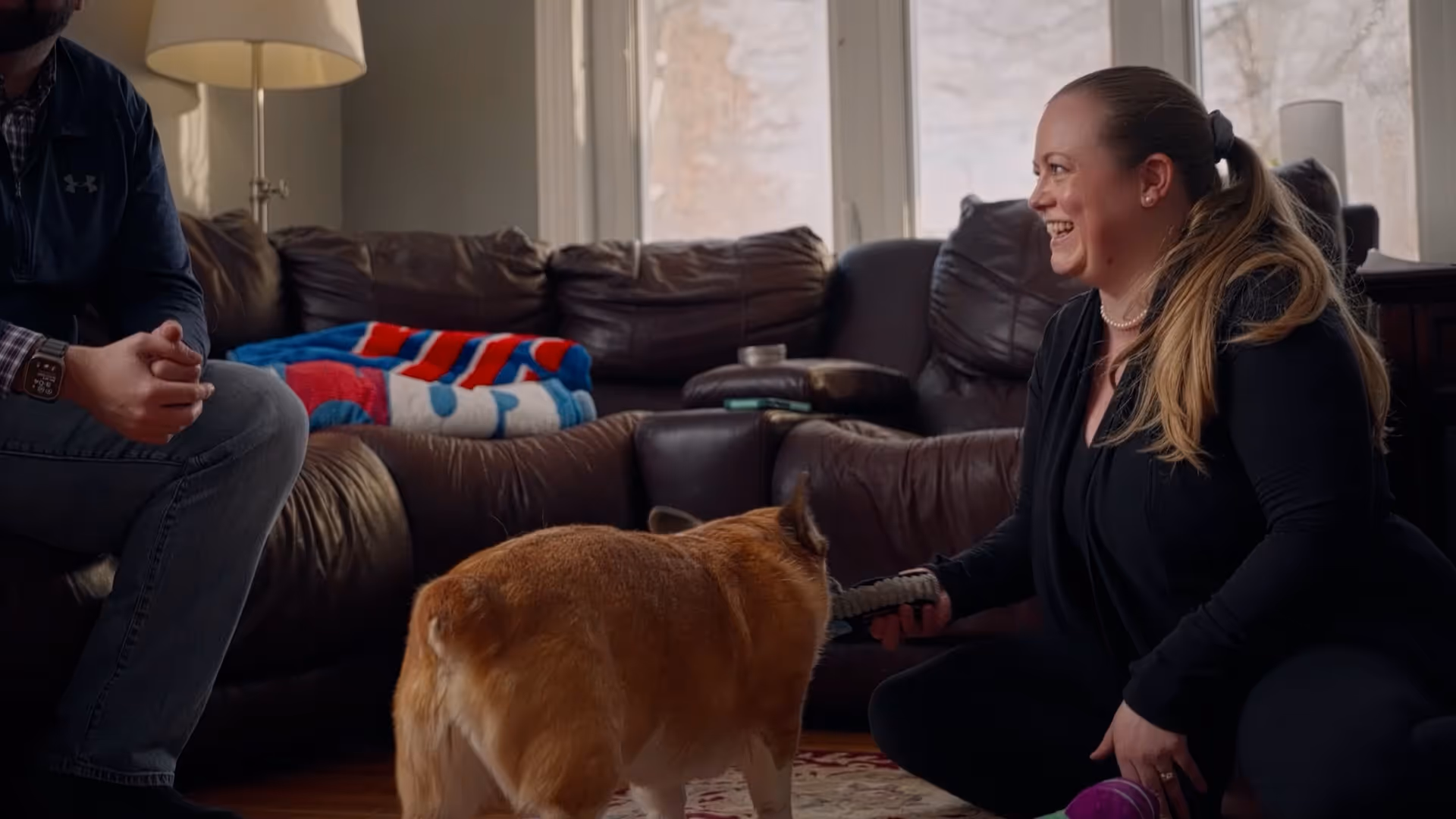 Woman sitting on floor playing with a dog in a living room with a couch and a lamp.