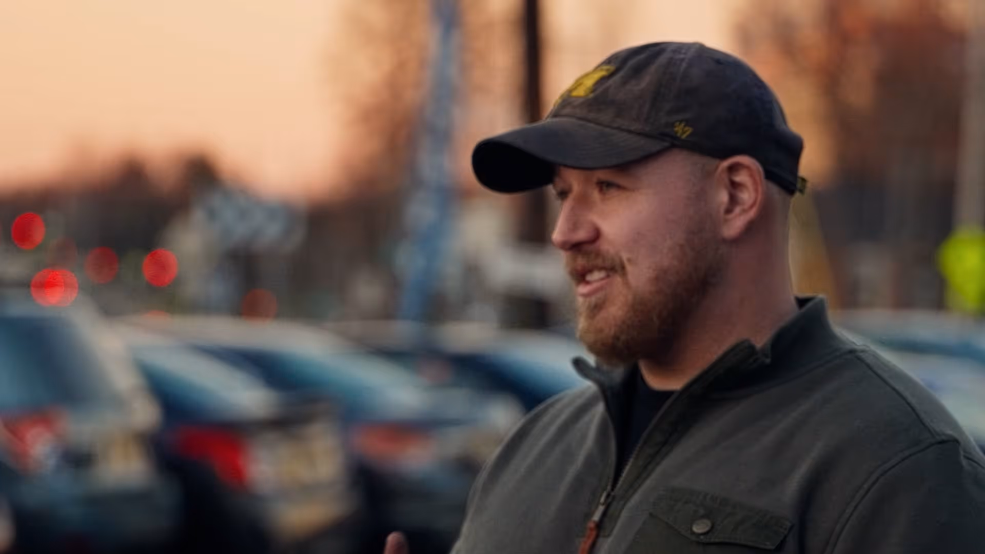 Bearded man wearing cap and green jacket standing with cars and sunset background.