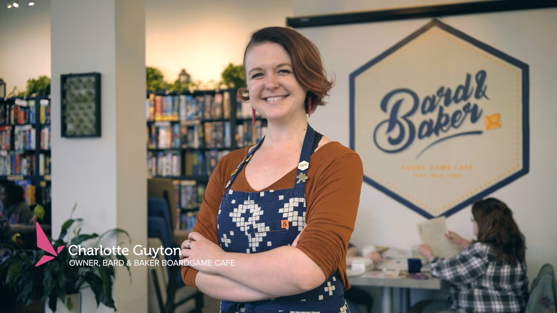 Smiling woman in an apron stands inside a boardgame café with shelves and players behind her.