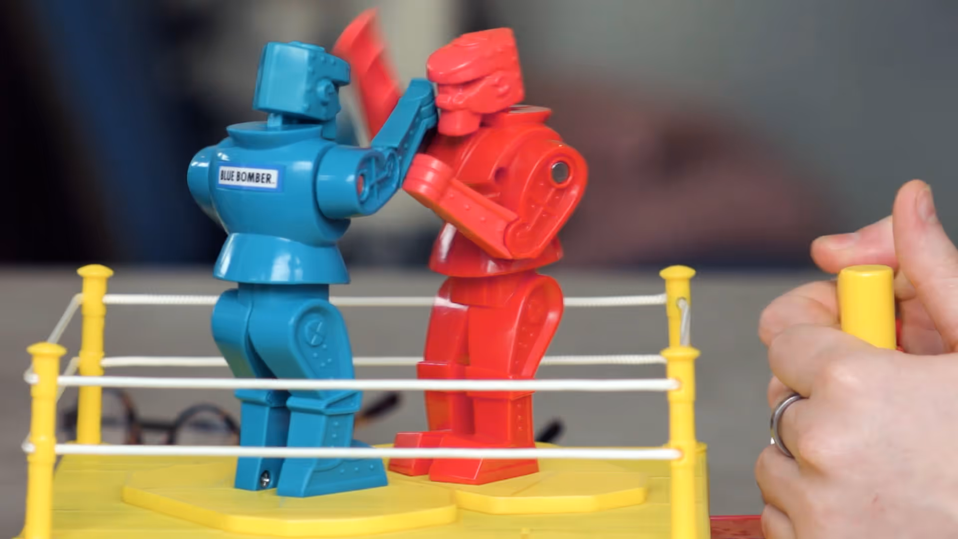 Blue and red toy robots boxing in a miniature ring with a hand controlling a lever.