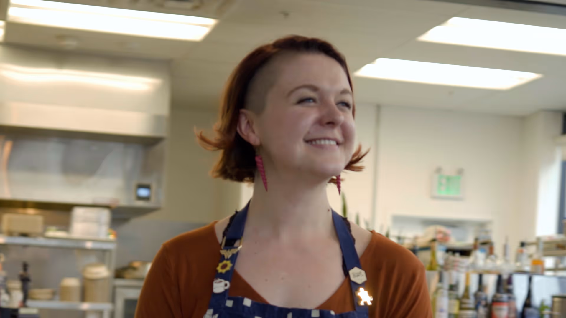 Smiling woman with short hair wearing apron and pink earrings in a kitchen setting.