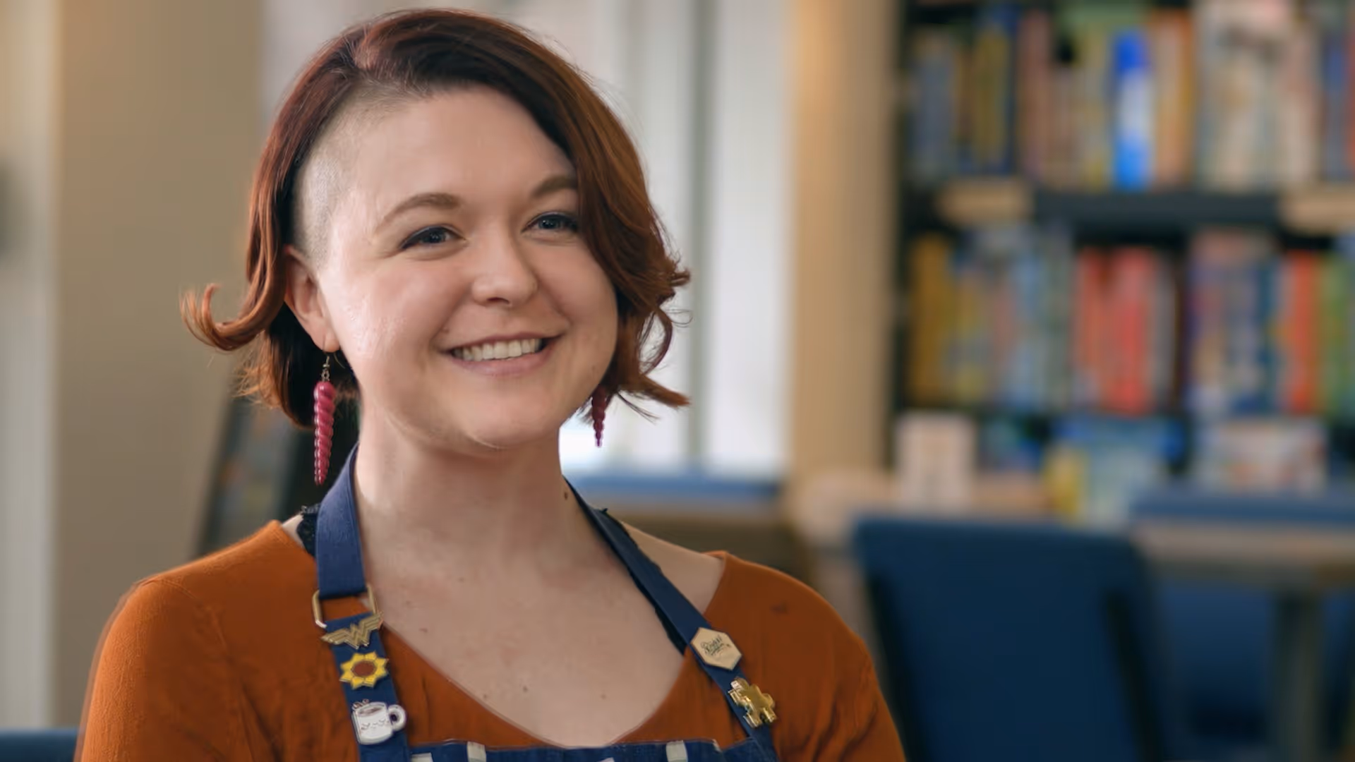 Smiling woman with short red hair, wearing spiral earrings and a badge-decorated apron indoors.