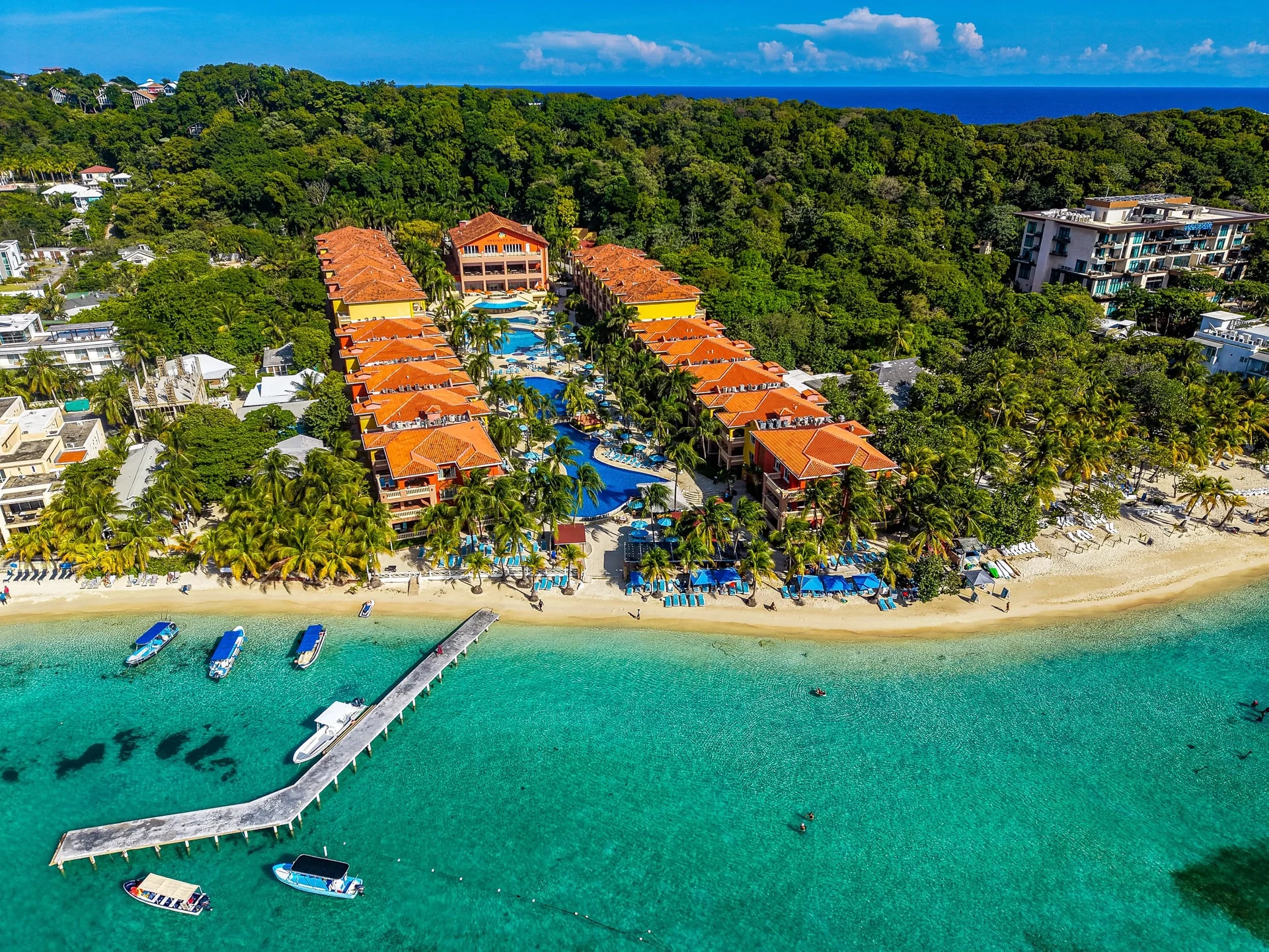 Aerial view of a tropical beach resort with orange-roofed buildings, turquoise shallow waters, boats, a pier, and lush green forest.
