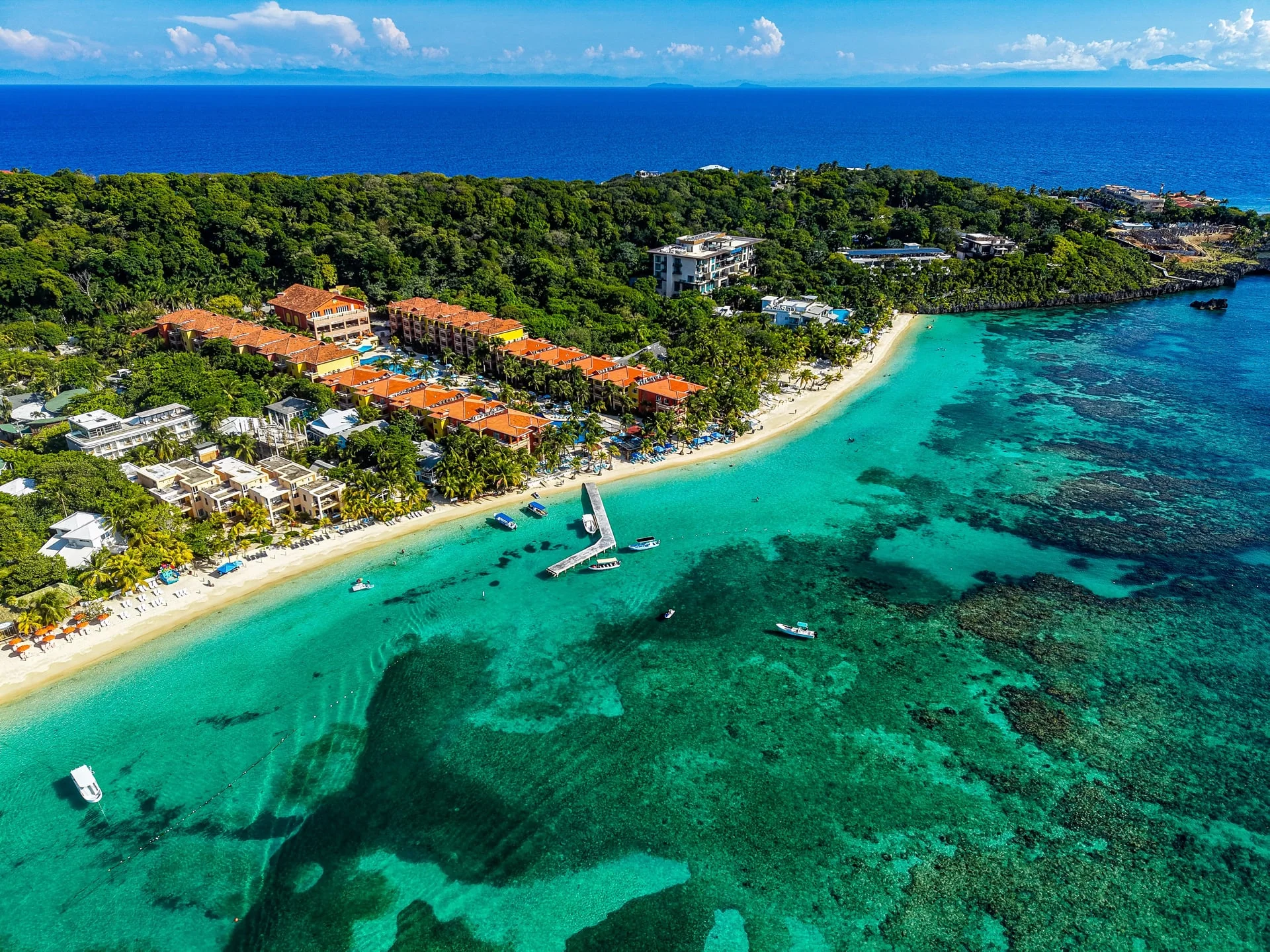 Aerial view of a tropical resort with orange-roofed buildings along a white sandy beach, surrounded by lush greenery and turquoise ocean with boats near a wooden pier.