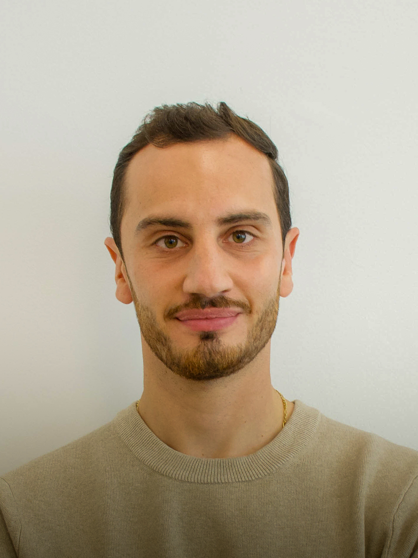 Portrait of a young man with short dark hair and a trimmed beard, wearing a black crewneck sweater against a plain gray background.