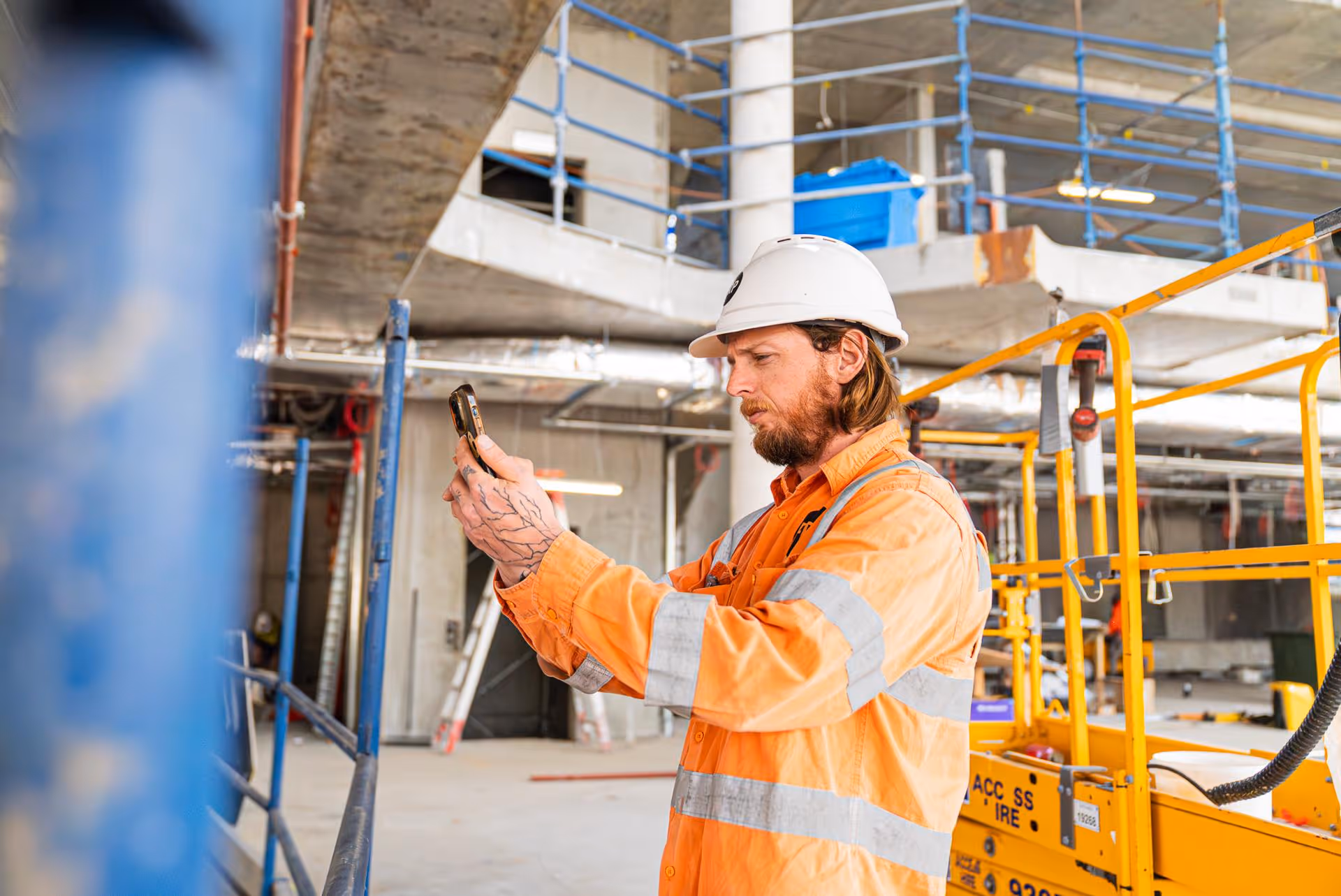 Construction worker in an orange safety jacket and white hard hat using a smartphone inside a building under construction.