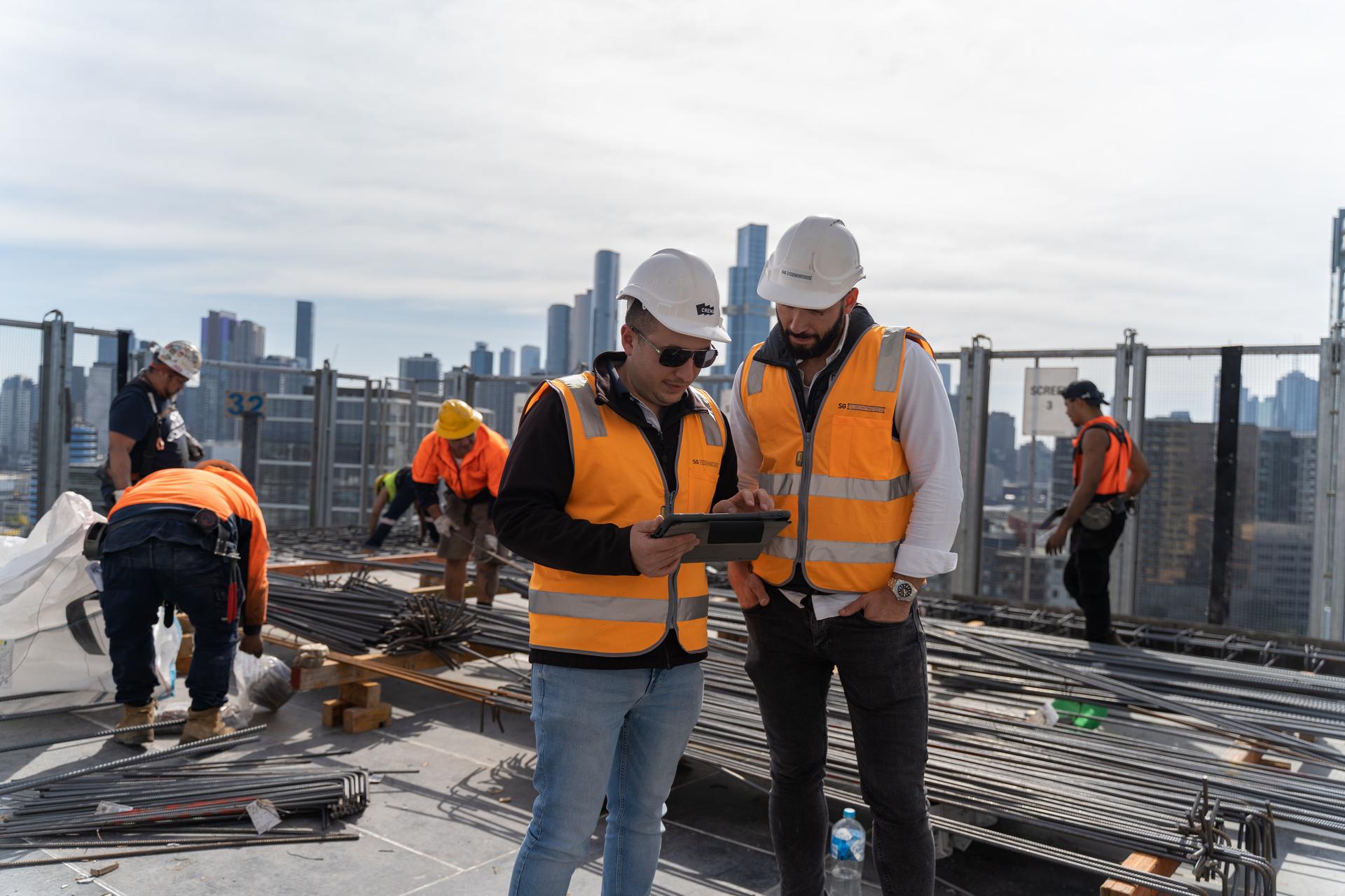 Two construction workers in orange safety vests and helmets reviewing information on a tablet at a building site with city skyline in the background.