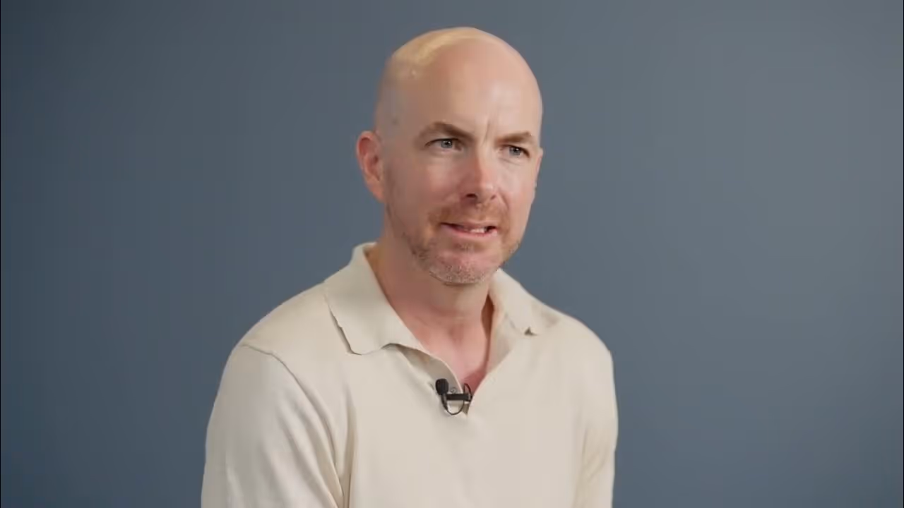 Bald man with light beard wearing a beige polo shirt speaking in front of a plain blue-gray background.