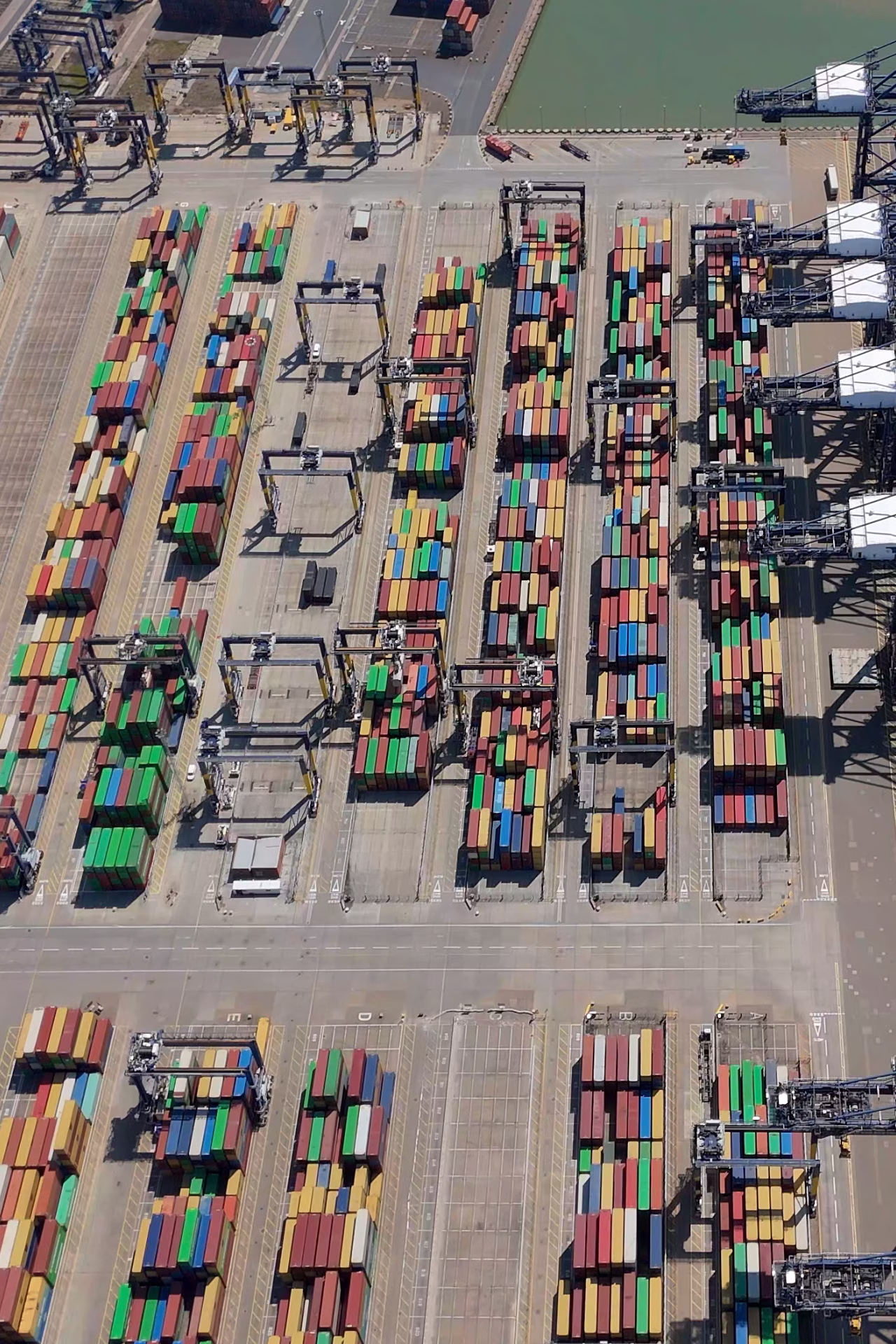 Vue aérienne d'un terminal portuaire avec de nombreuses rangées de conteneurs colorés empilés et des grues de chargement.