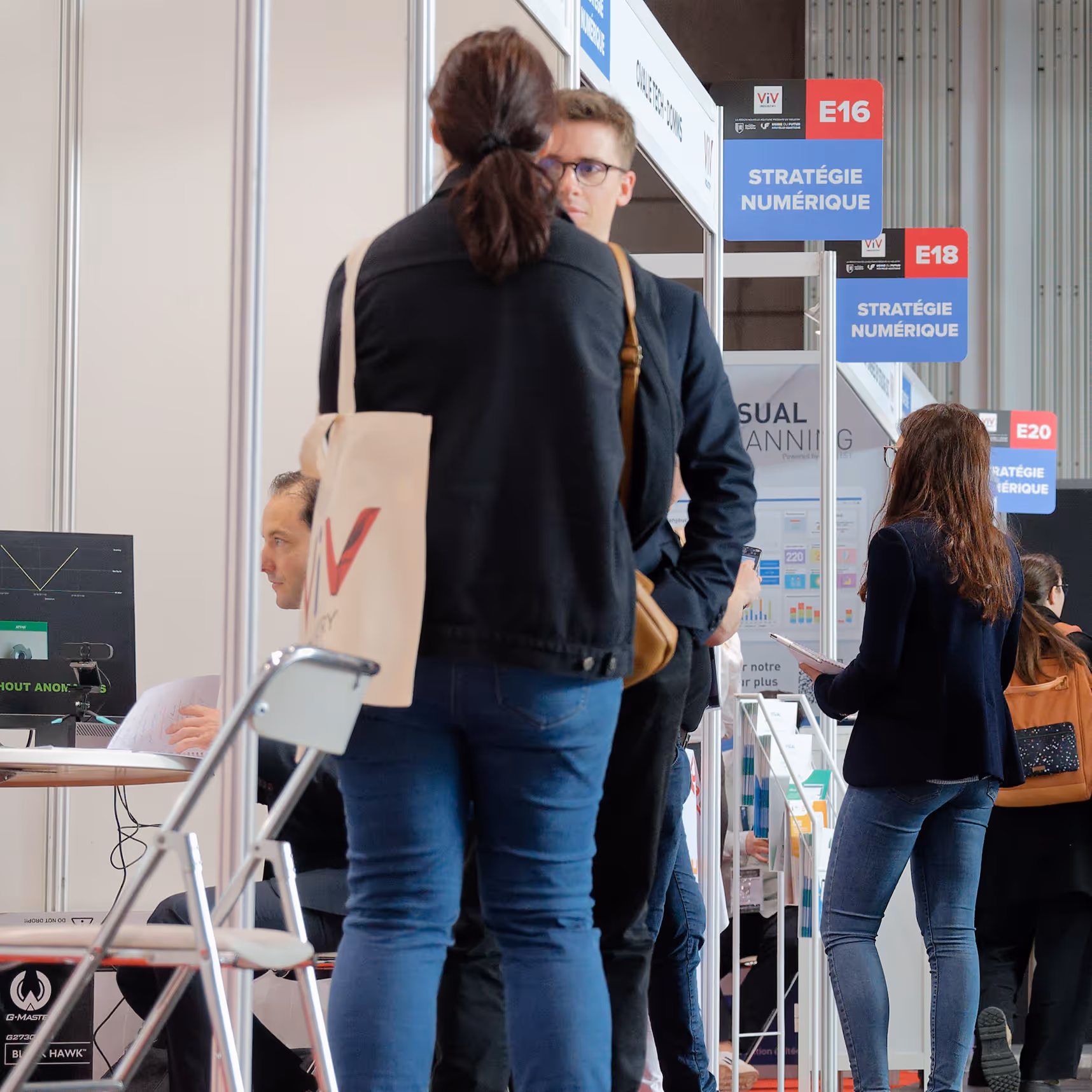 Personnes échangeant dans un stand d'exposition avec des panneaux indiquant "Stratégie numérique" aux emplacements E16, E18 et E20.