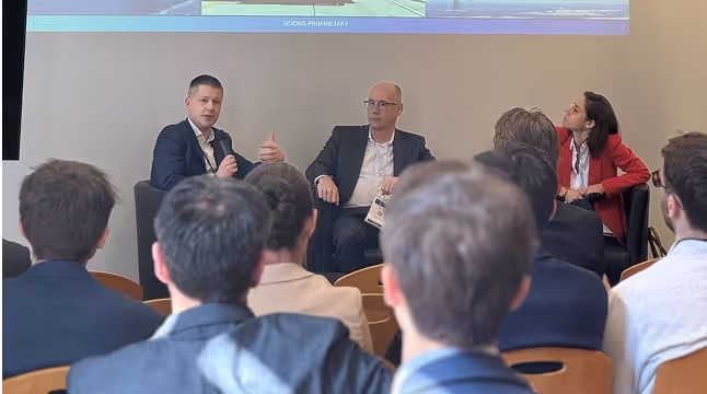 Trois intervenants assis devant un public lors d'une conférence, avec un homme à gauche parlant dans un micro.