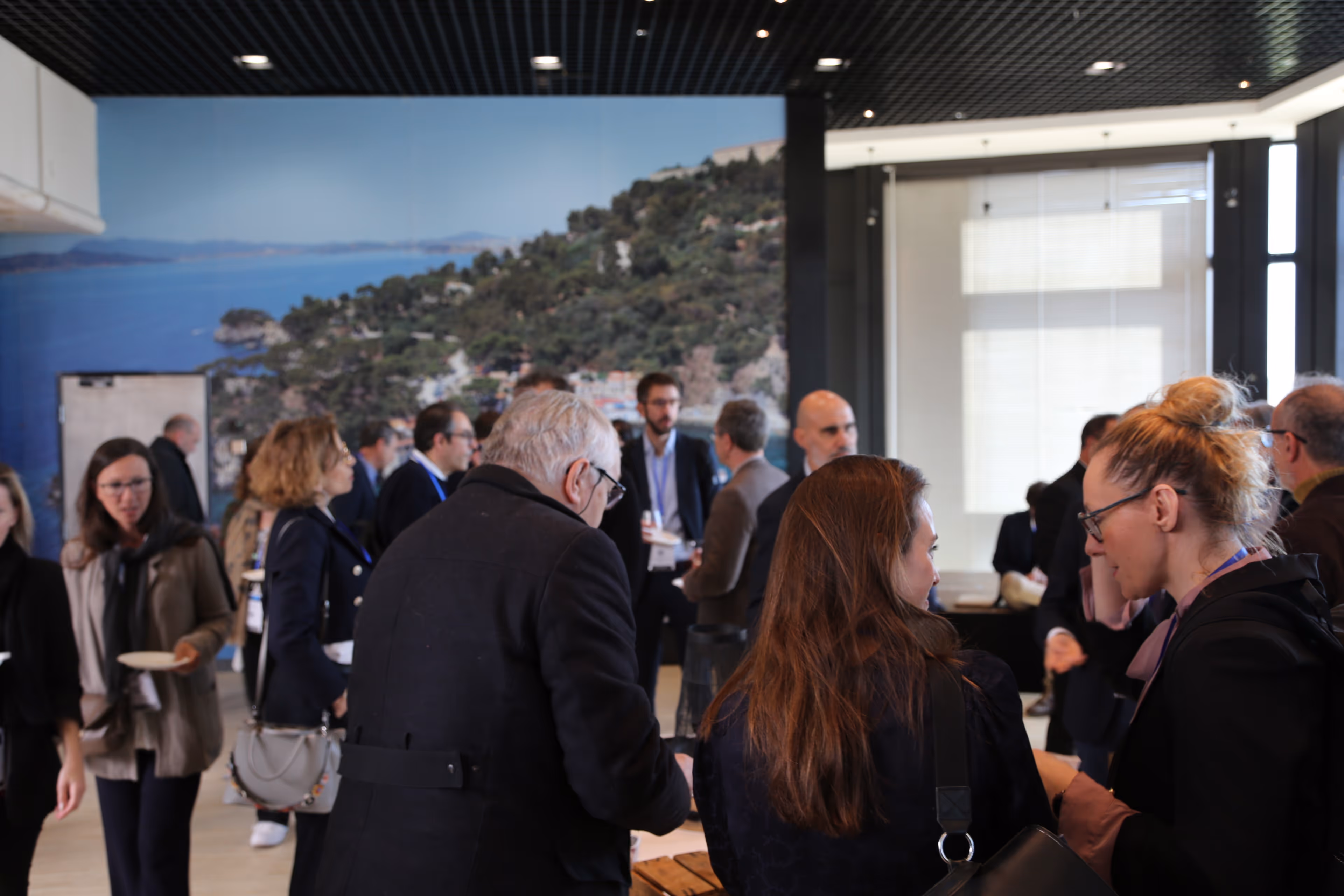 Un groupe de personnes discutant lors d'un événement en intérieur avec une grande photo murale d'un paysage côtier en arrière-plan.