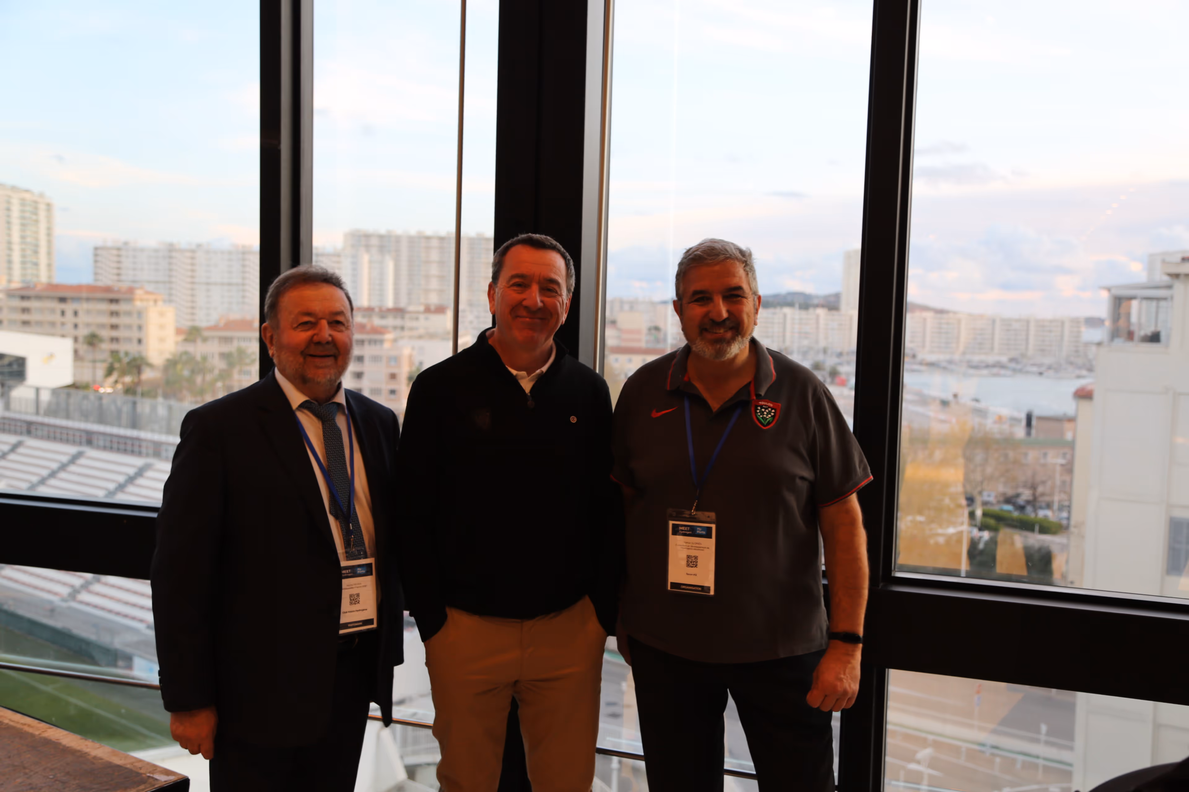 Trois hommes souriants portant des badges d'identification posant devant une grande fenêtre avec vue sur des immeubles et un port.
