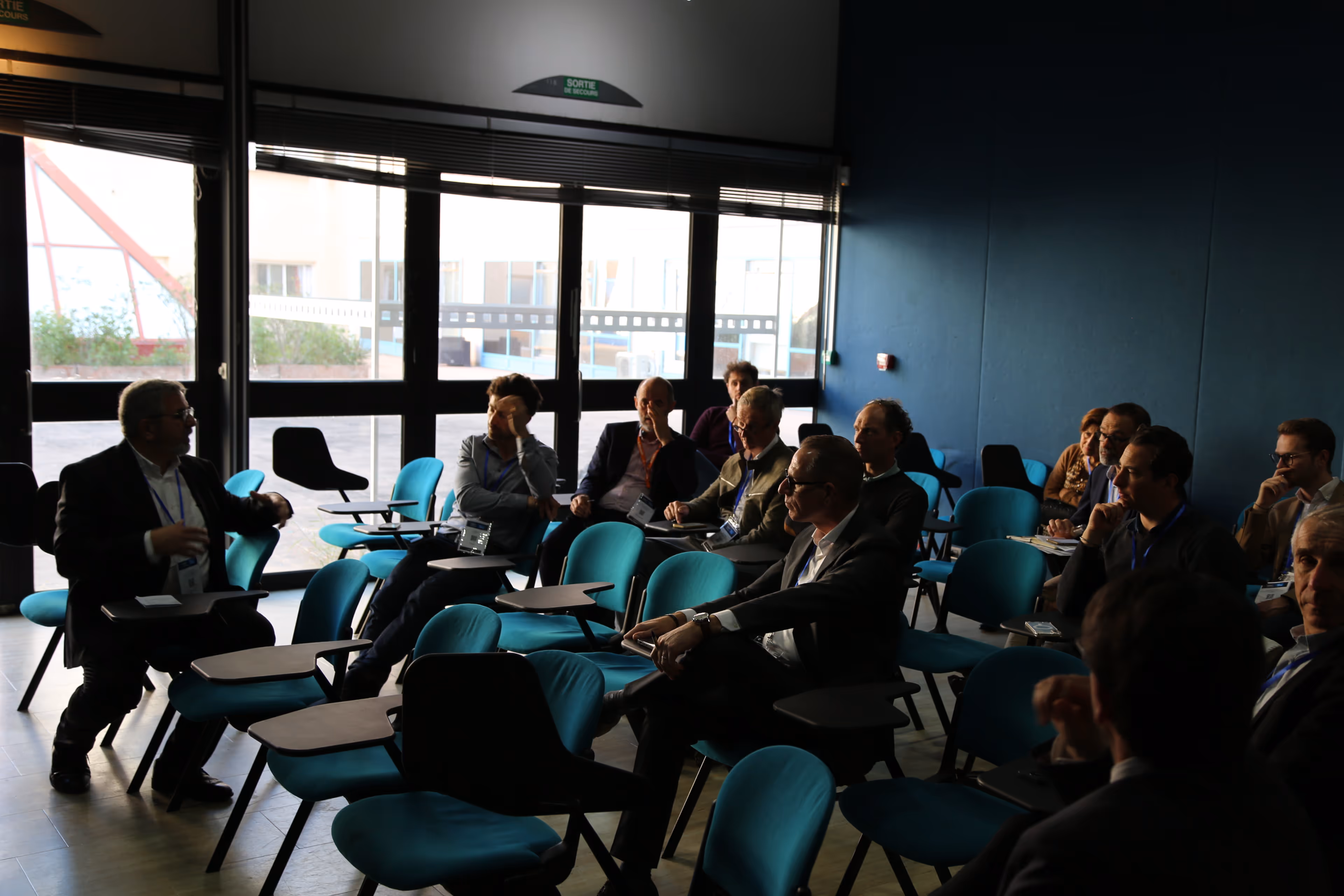 Groupe de personnes en réunion assises sur des chaises bleues avec tablettes dans une salle sombre près d'une grande fenêtre.