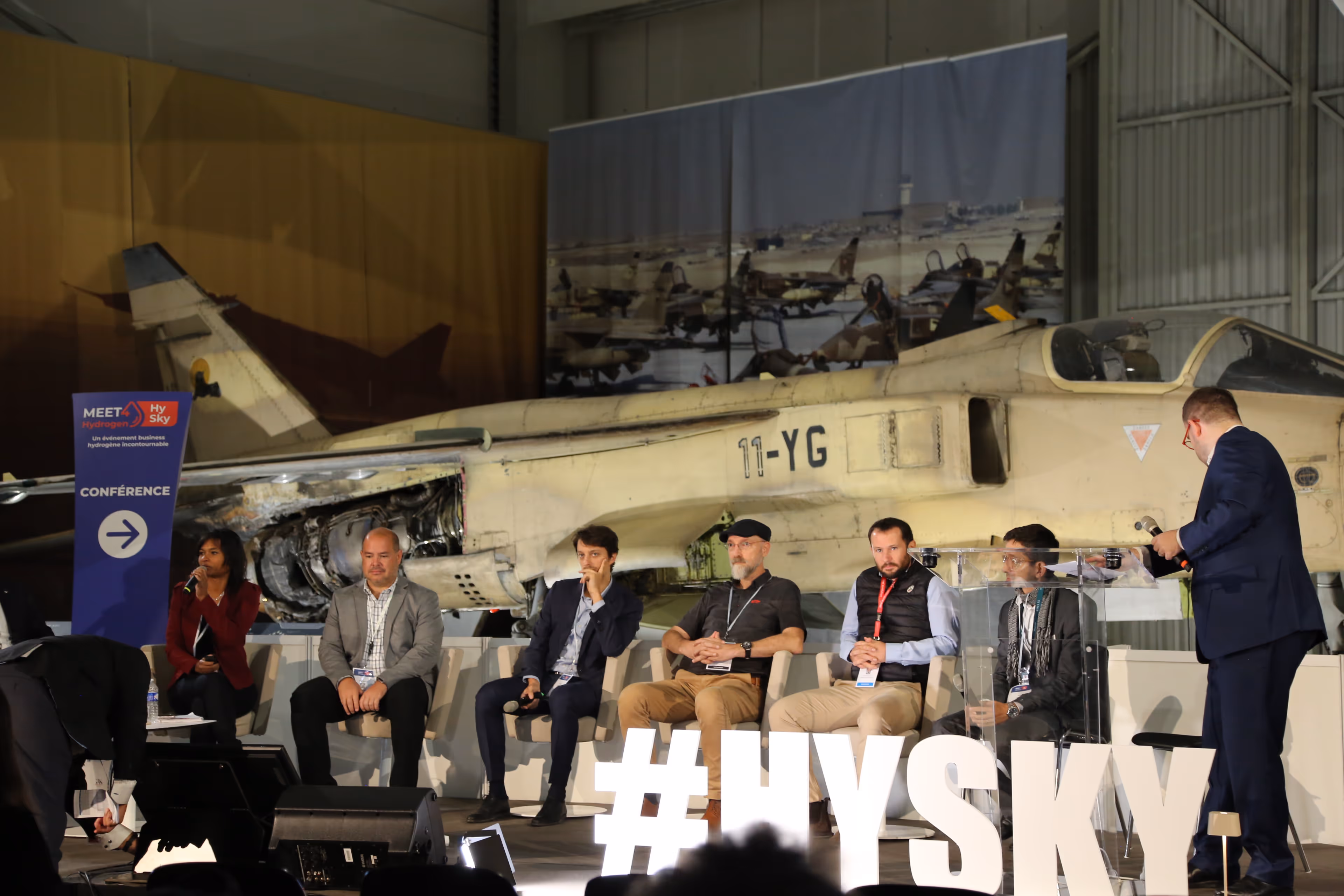 Un panel de six intervenants assis devant un avion de chasse militaire beige lors d'une conférence HySky sur l'hydrogène.