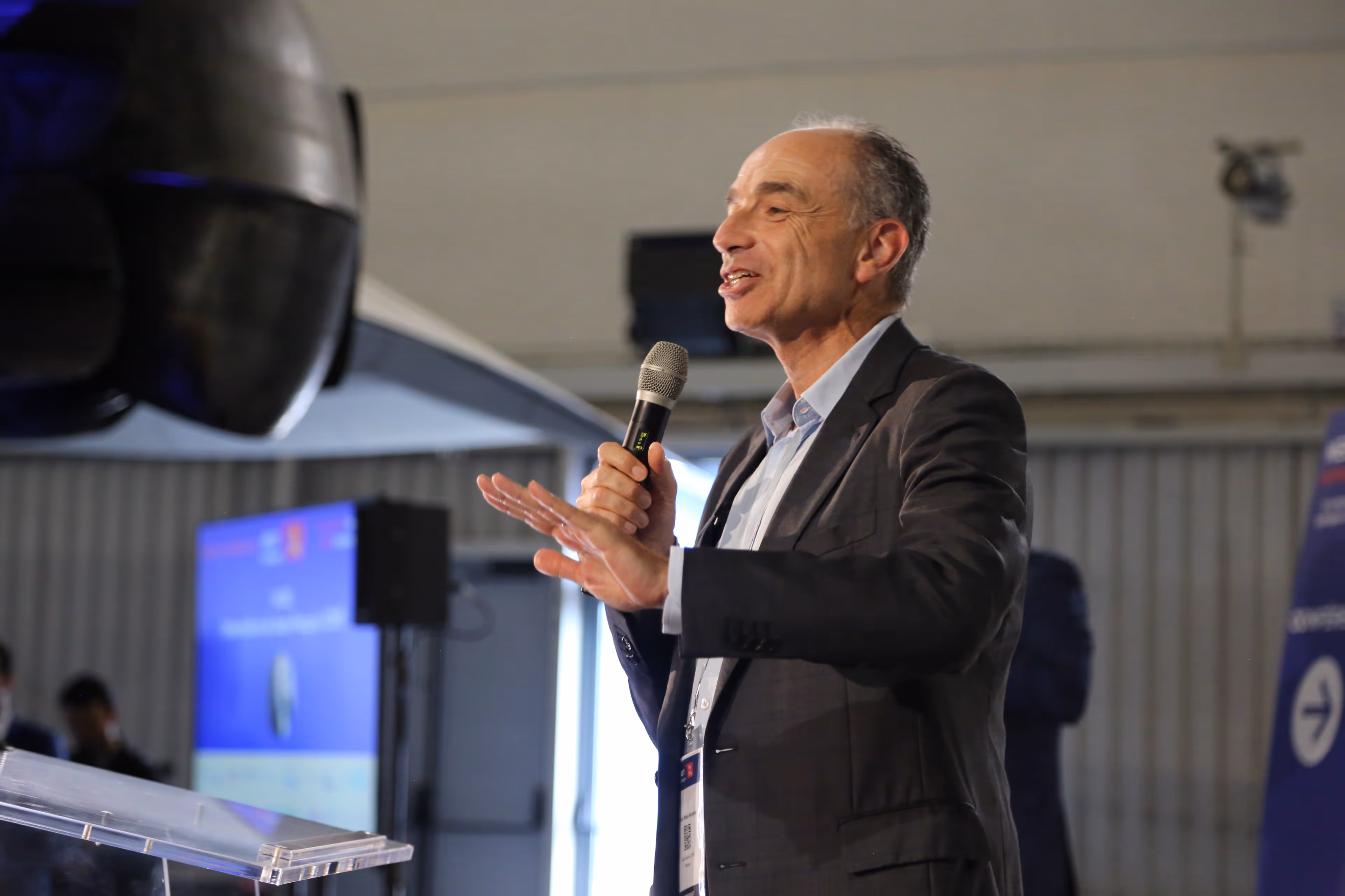 Homme d'âge moyen en costume parlant au micro lors d'une conférence en intérieur.