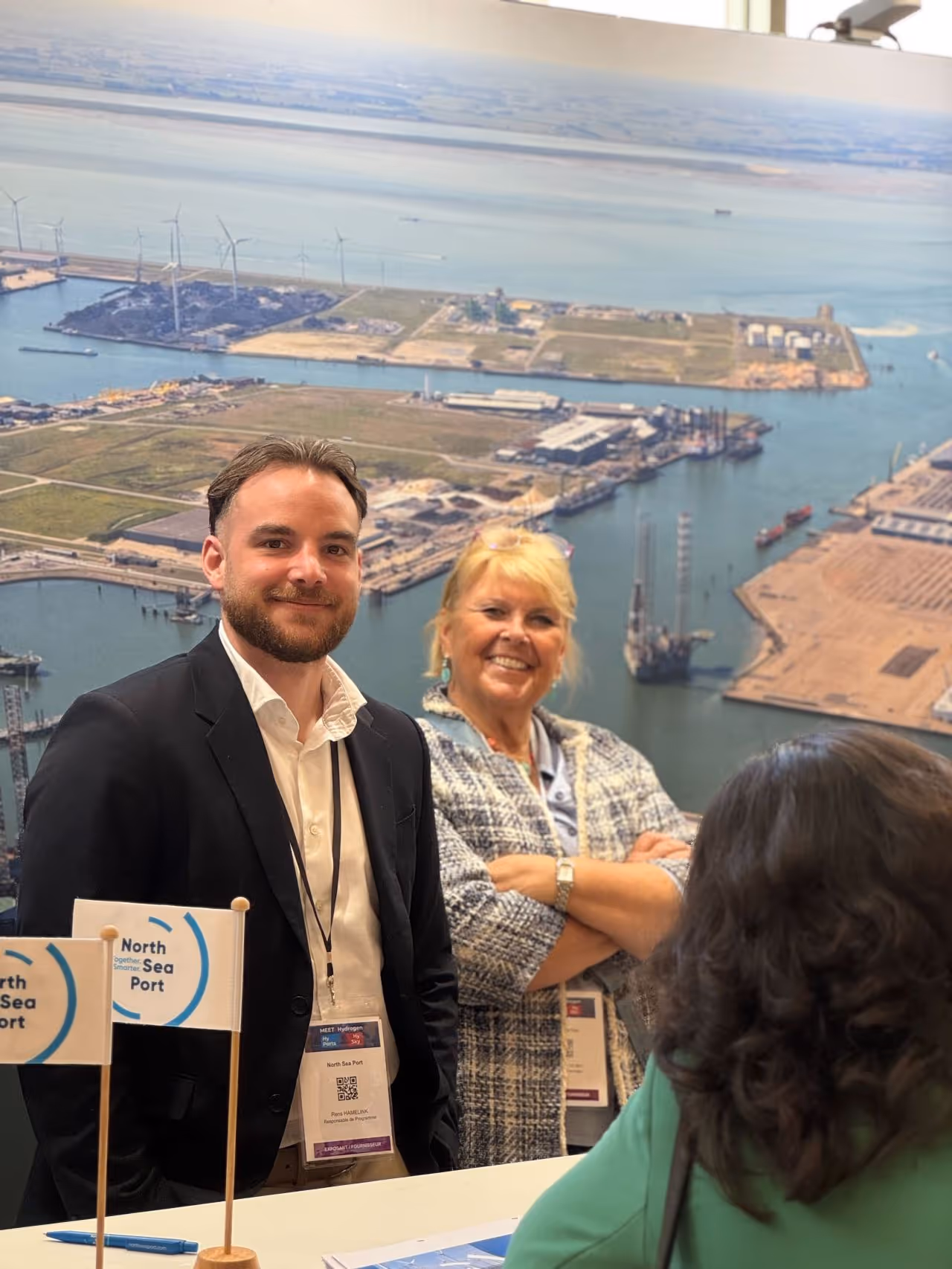 Deux professionnels souriants debout devant une grande photo aérienne d'un port maritime, discutant avec une femme de dos.