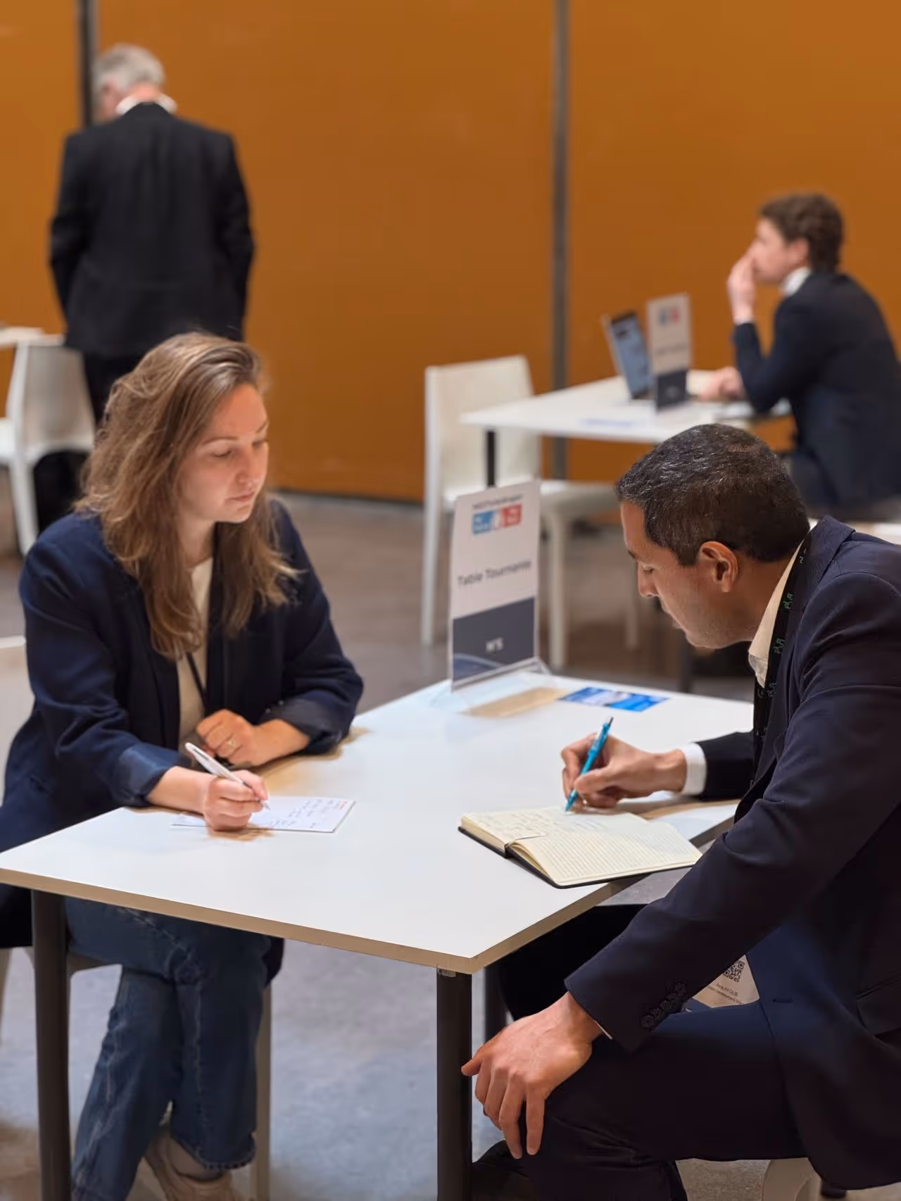Une femme et un homme en costume assis à une table blanche, écrivant dans un carnet et sur une feuille lors d'une réunion professionnelle.