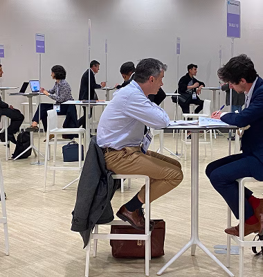 Deux hommes discutent assis à une table haute dans une salle lumineuse avec d'autres personnes en arrière-plan lors d'un événement professionnel.