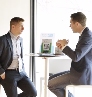 Deux hommes en costume discutant assis à une table haute près d'une fenêtre dans un environnement lumineux.