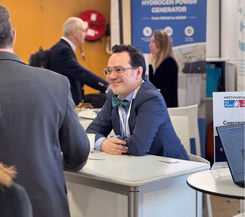 Un homme souriant avec un nœud papillon et un badge de conférence assis à un stand lors d'un événement professionnel.