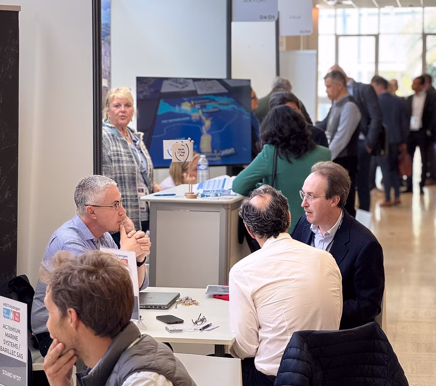 Personnes en réunion autour d'une table lors d'un salon professionnel animé, avec un écran et plusieurs participants en arrière-plan.