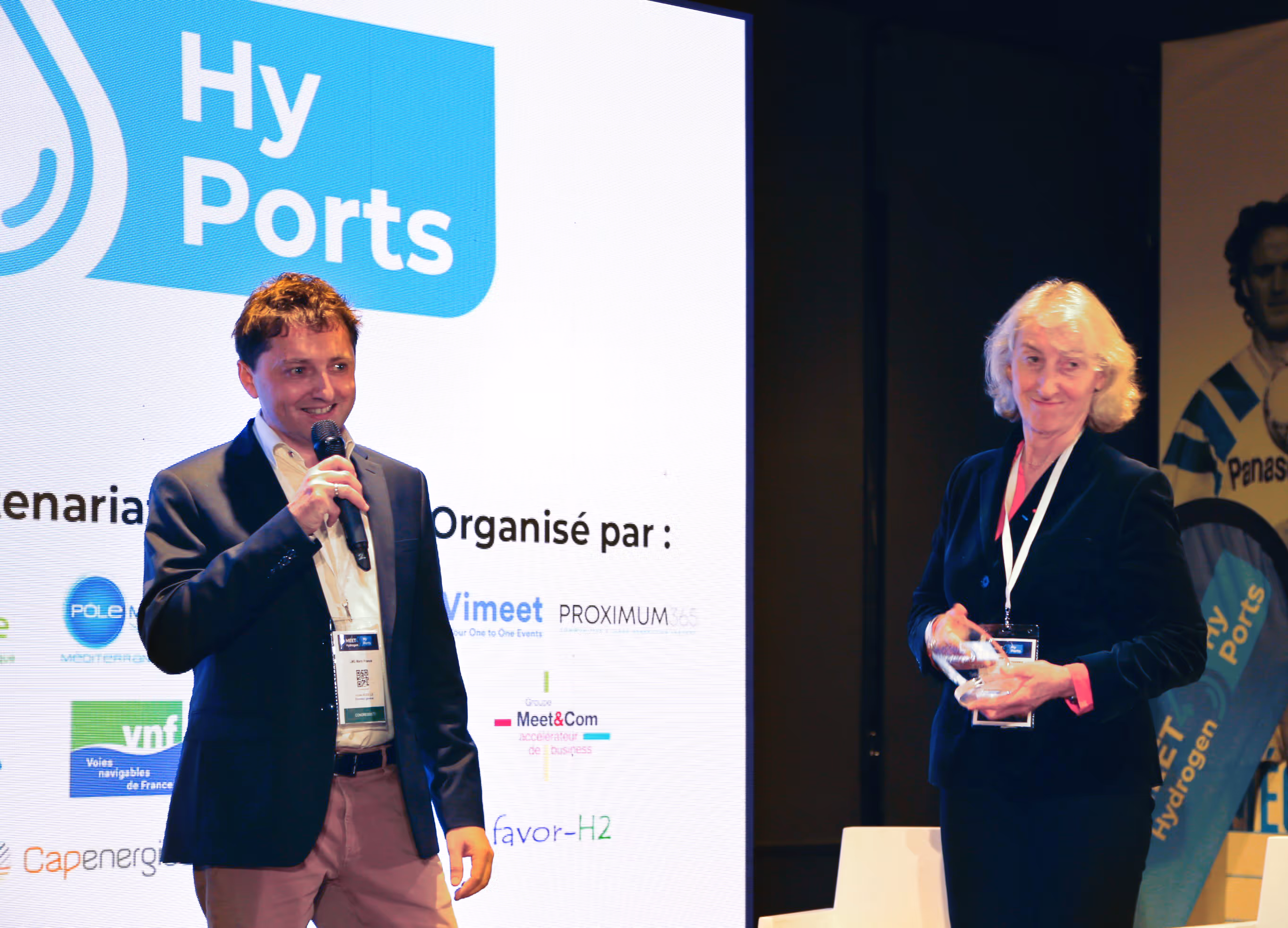 Un homme tenant un micro en costume sourit devant un écran portant le logo Hy Ports, tandis qu'une femme avec un badge tient un trophée près de lui.
