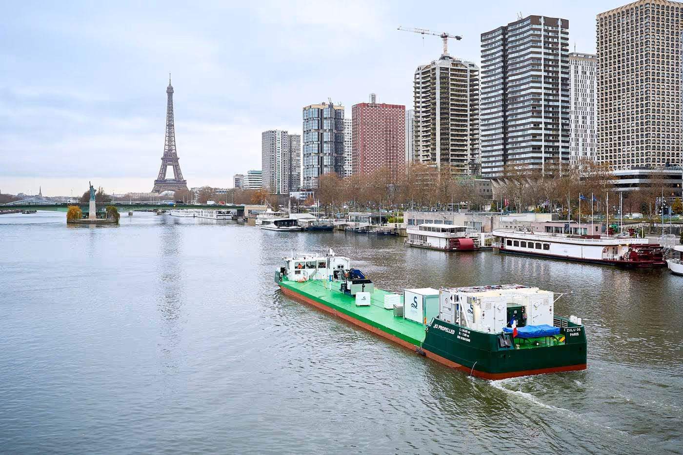 Bateau cargo vert naviguant sur la Seine avec la Tour Eiffel et des immeubles en arrière-plan à Paris.