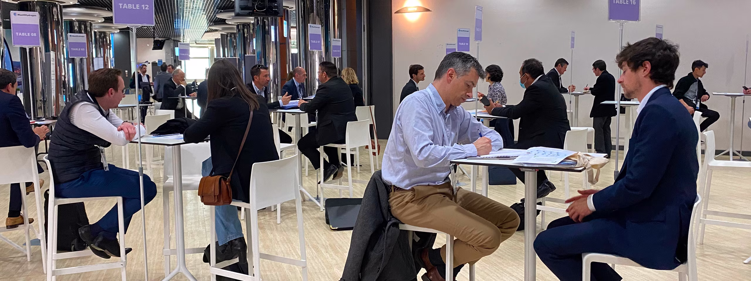 Plusieurs personnes en réunion assises à des tables hautes dans une salle de conférence lumineuse, échangeant et prenant des notes.