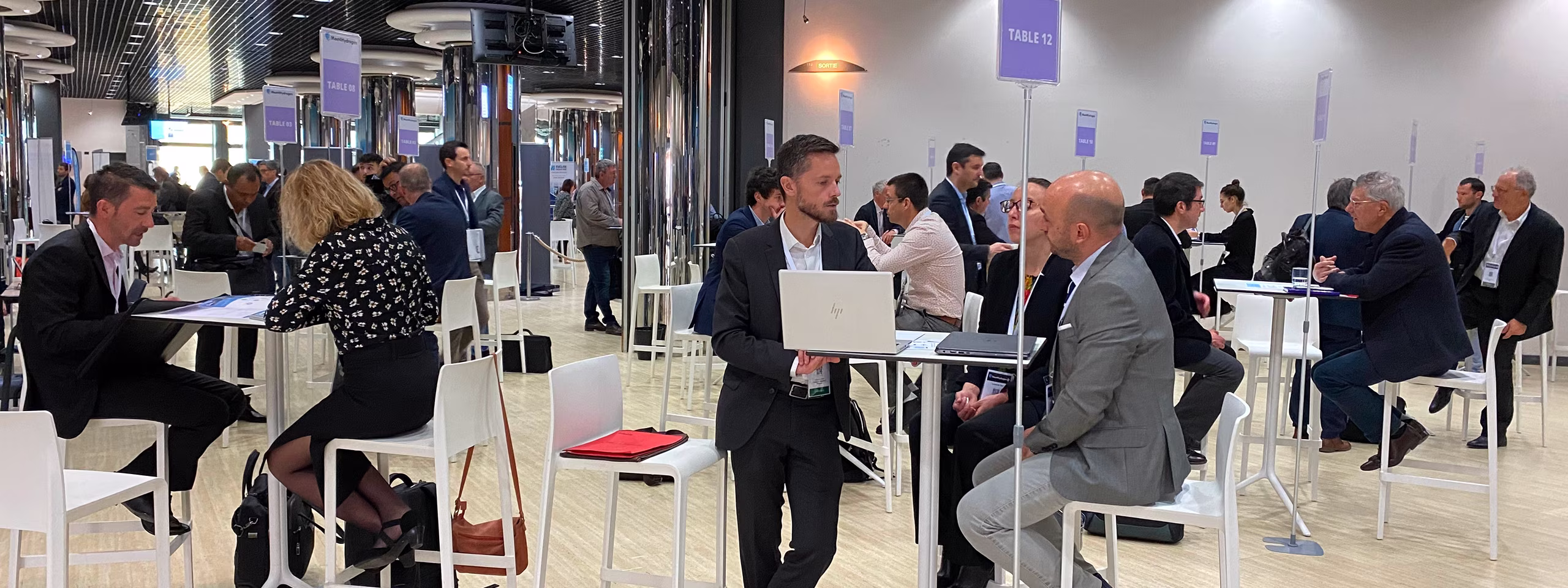 Personnes discutant en petits groupes assis à des tables hautes dans une salle de conférence moderne.