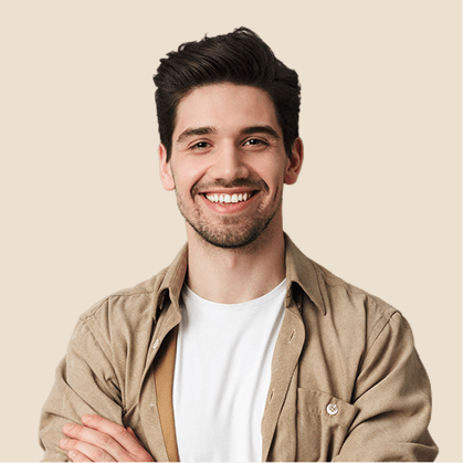 Smiling young man with dark hair wearing a beige shirt over a white t-shirt, standing with arms crossed against a light beige background.