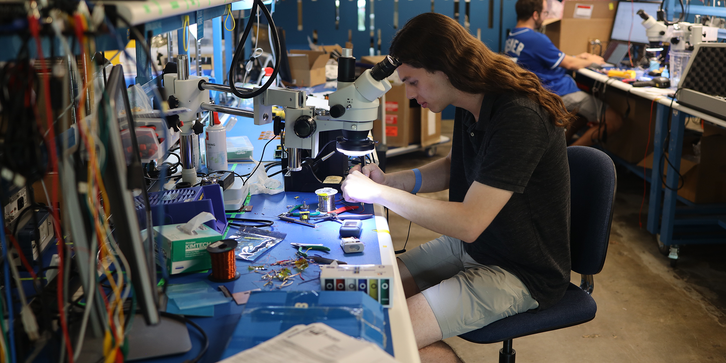 Young man with long hair working on electronic components under a microscope at a cluttered workbench in a lab.