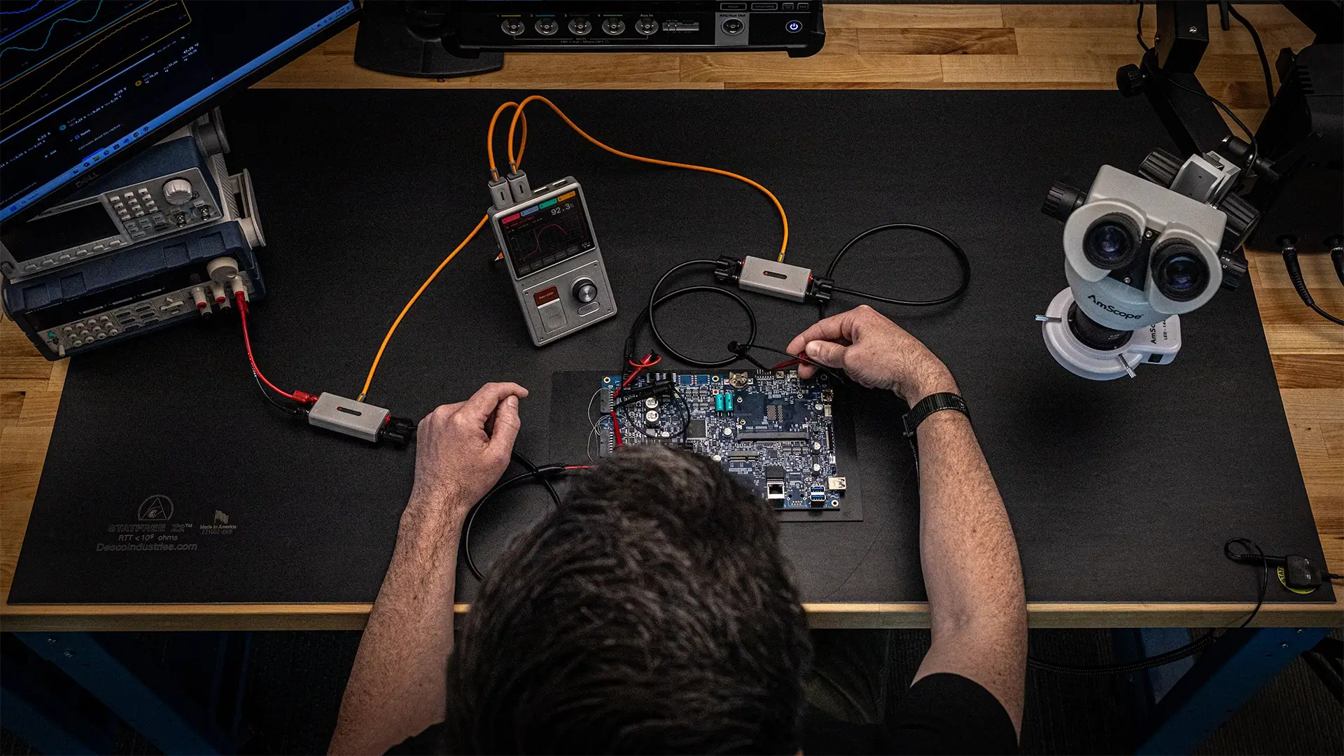 Person testing a circuit board on a workbench with electronic measuring equipment and a microscope.