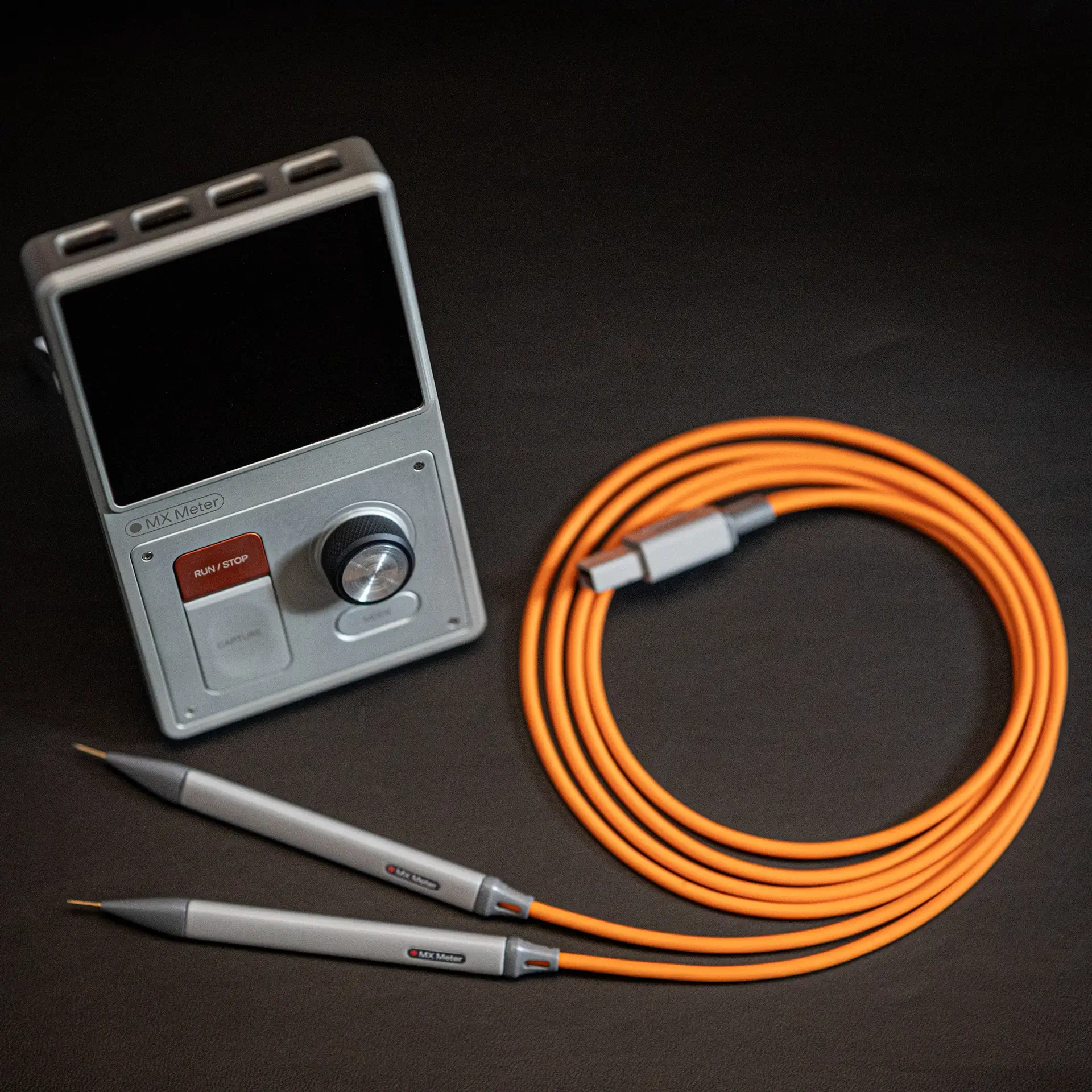 MX Meter device with a color display showing waveform graphs and data, connected to two white pen-shaped probes with orange cables.