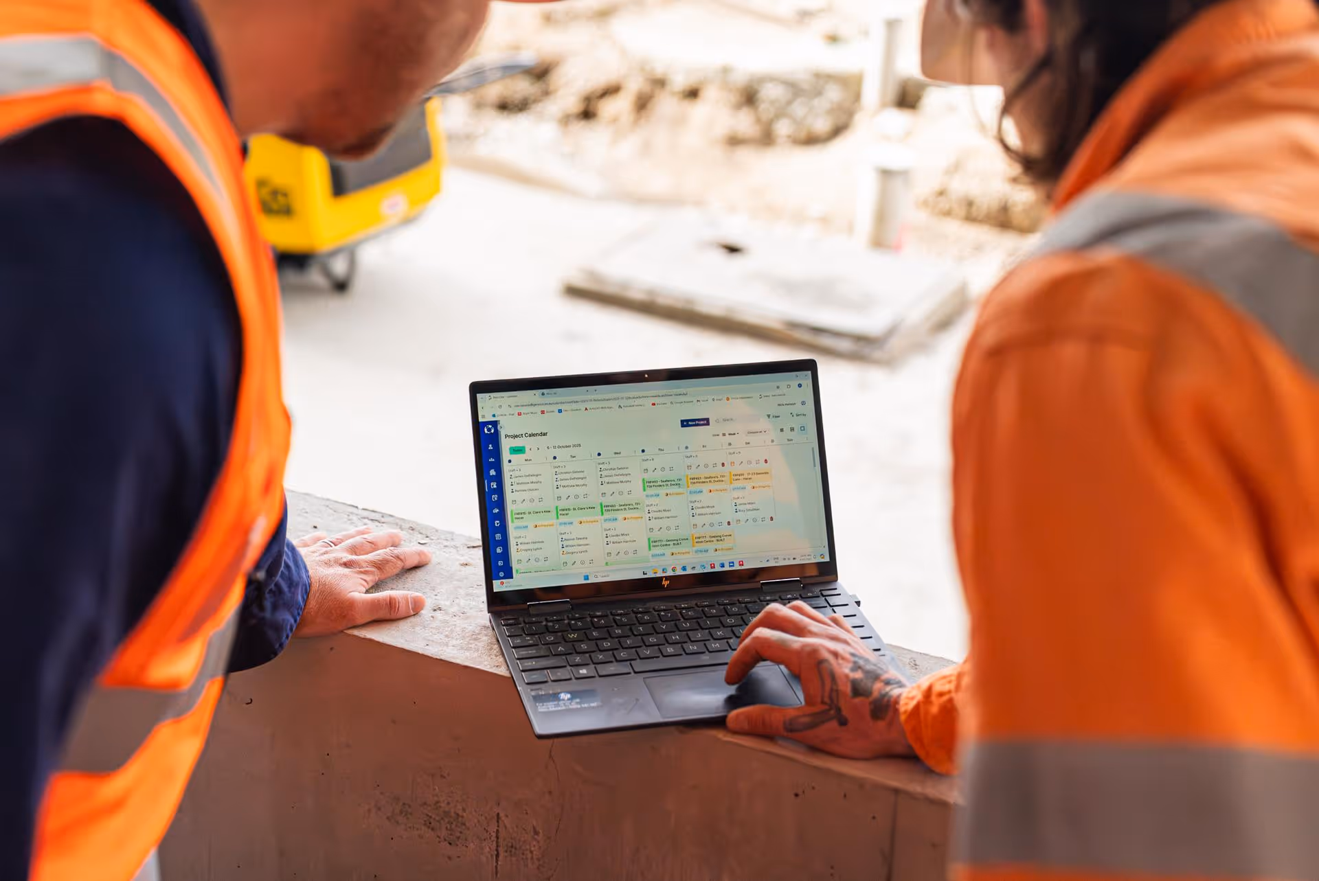 Two workers in hi-viz clothing, looking at the Neo Intelligence Project Calendar on a laptop whilst being on site.