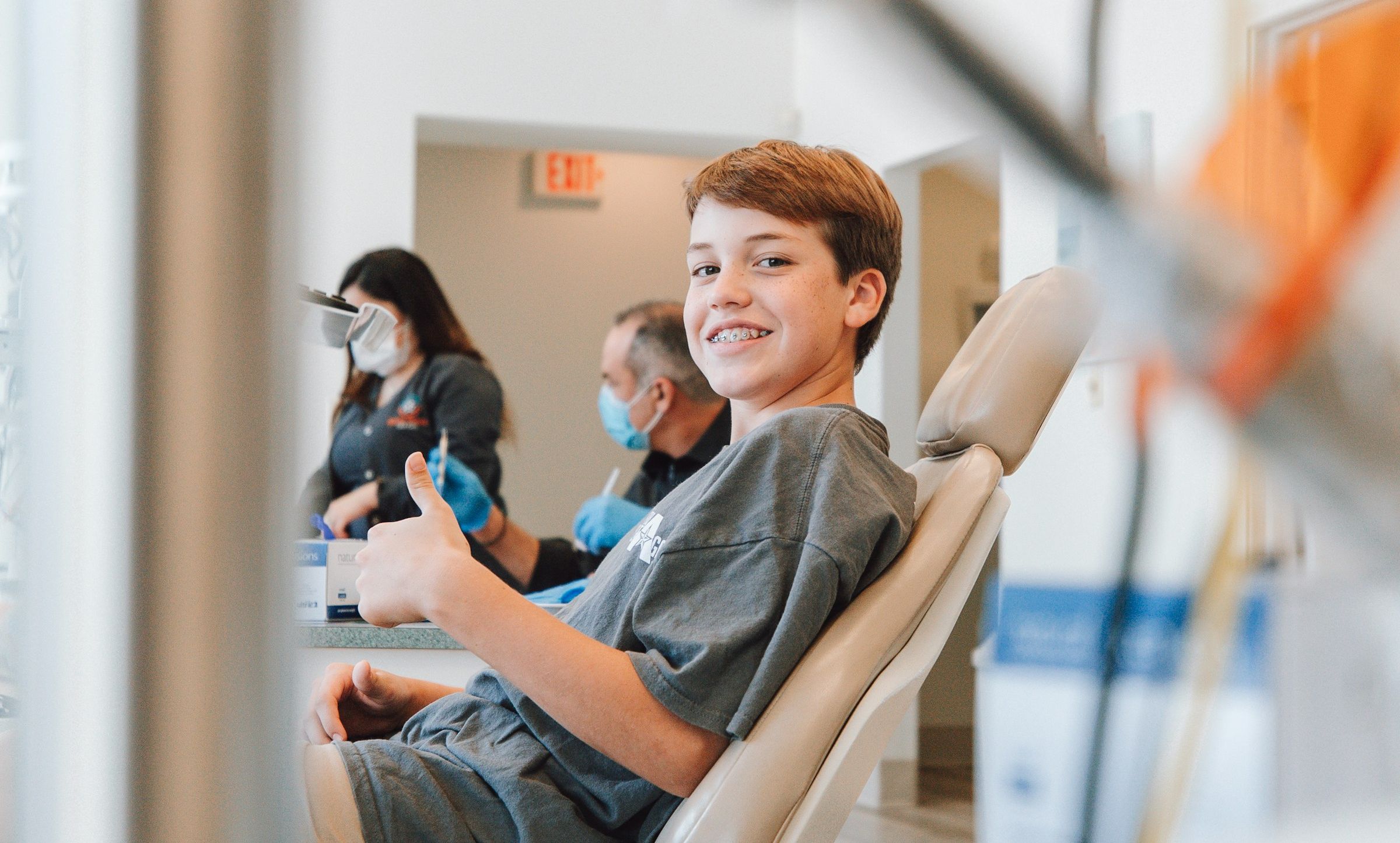 Smiling boy with braces sitting in a dental chair giving a thumbs up, with dental professionals wearing masks in the background.