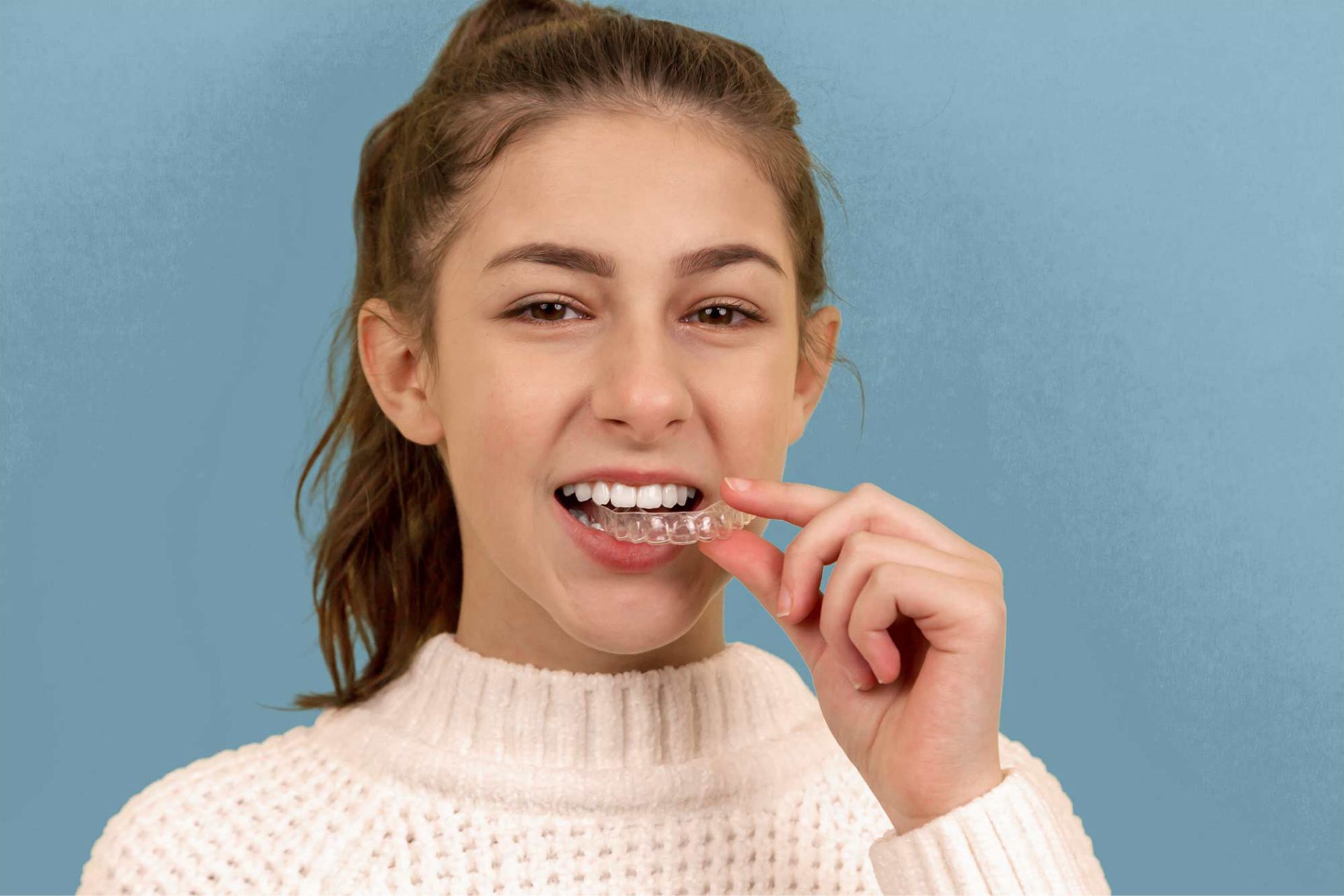 Young woman wearing a white sweater is holding a clear dental aligner near her mouth.