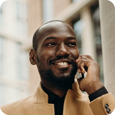 Smiling man in tan jacket talking on a phone with blurred urban background.