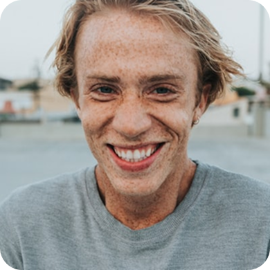 Smiling young person with light skin, freckles, and blond hair wearing a gray shirt outdoors.